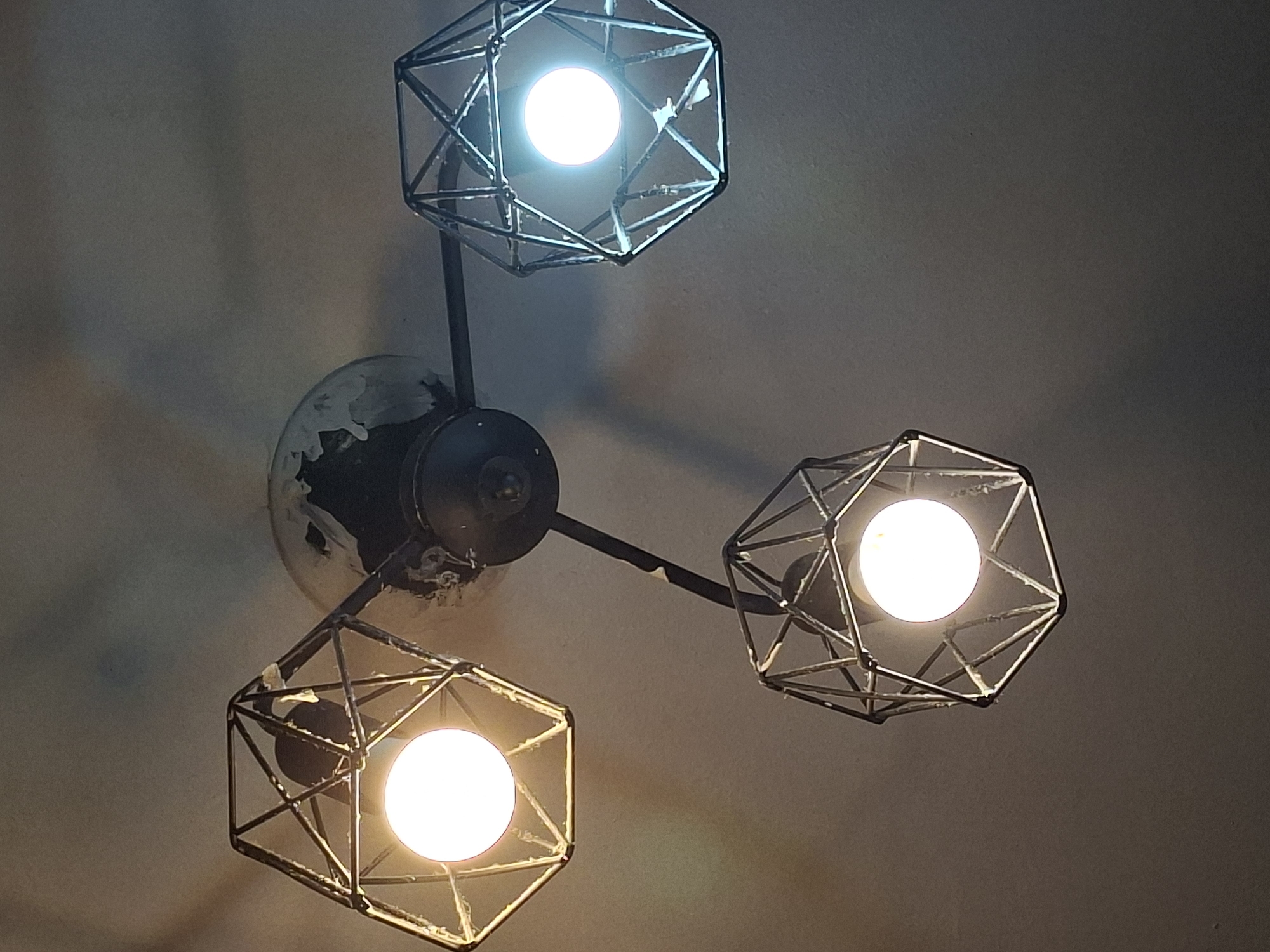 The image shows a close-up, low-angle view of a three-arm ceiling light fixture. Each arm extends from a central, dark-colored mounting base attached to a textured, off-white ceiling. The mounting base has some visible wear and tear, with what appears to be white residue or debris around its edges. Each arm supports a geometrically shaped lampshade, constructed from dark metal bars forming a polyhedral shape. Inside each shade, a round, white light bulb is illuminated. The bulb on the left emits a warm, yellow-toned light, while the bulb on the right and the bottom bulb emit a cooler, bluish-white light. The overall lighting in the image is dim, with the light from the bulbs creating bright spots and casting shadows on the ceiling. The image does not contain any elements that would allow for the identification of a specific location in Moscow, Russia, or any other city.
