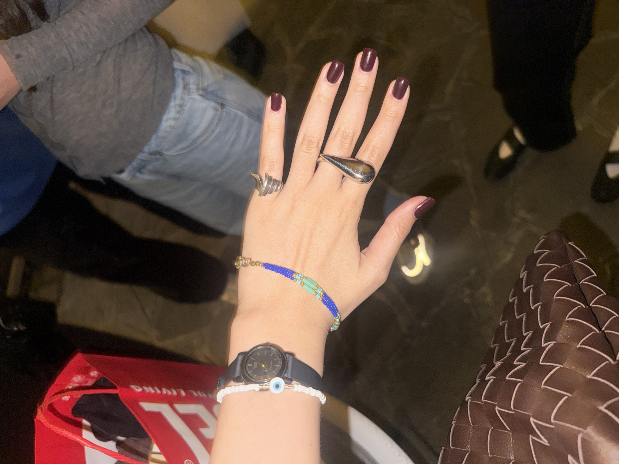 A hand with burgundy nails adorned with two silver rings is prominently displayed. The hand wears a blue and gold beaded bracelet, a black wristwatch, and a white beaded bracelet with a blue eye charm. The arm is visible with gray clothing, while the body appears to wear denim jeans. A red shopping bag with white lettering is visible nearby, as well as a woven brown purse. In the background, legs in dark clothing and dark shoes are observed, and the floor appears to be dark, possibly marble or stone. The photo appears to be taken indoors. Without additional elements, it is impossible to pinpoint the exact location in Milan.