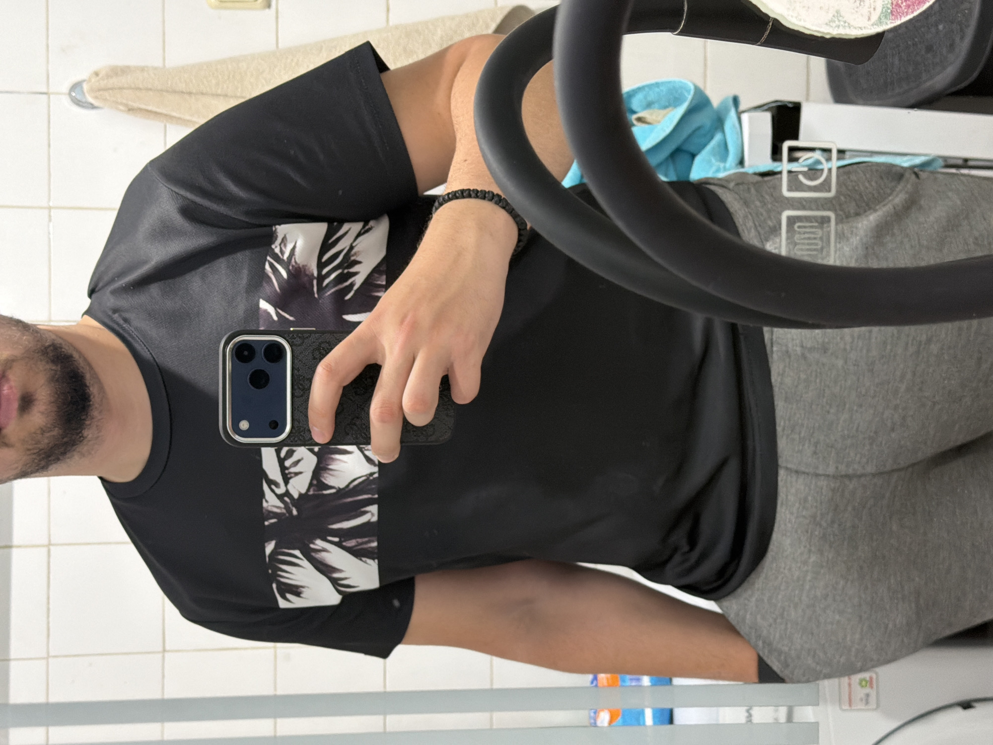 A man is taking a selfie in a bathroom or laundry room with a white tiled wall. He is wearing a black t-shirt with a white palm tree design on it, and grey sweatpants. He is holding a black smartphone with a silver case, taking a selfie in a mirror. The man has a beard and is wearing a black beaded bracelet on his left wrist. In the background, there is a towel rack with a beige towel, a blue towel, and what appears to be the black arm of an elliptical exercise machine. The bottom of a white appliance, possibly a washing machine or dryer, is also visible. The location in the city of Burgas, Bulgaria is unknown.