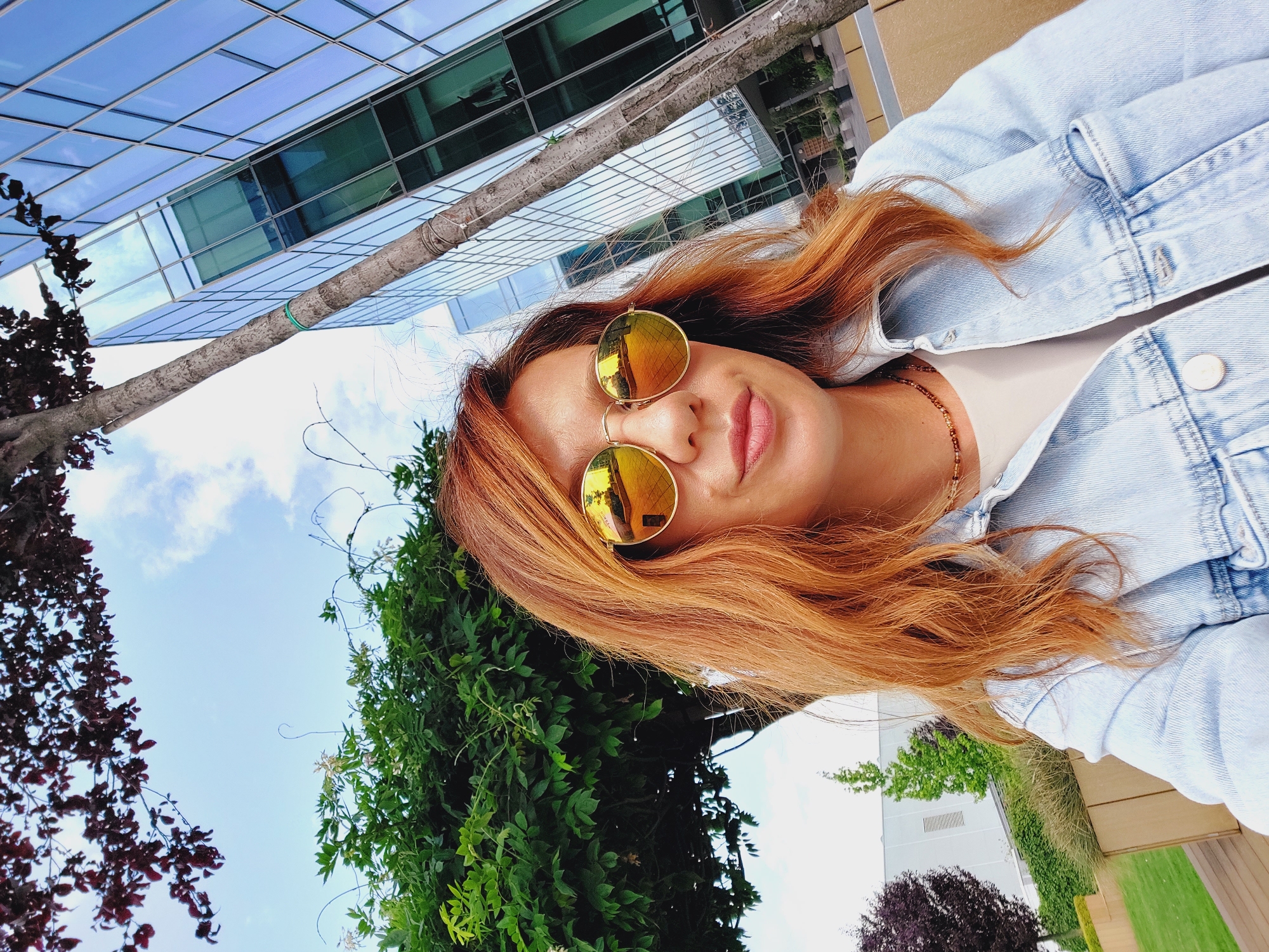 A woman with auburn hair is taking a selfie outdoors. She is wearing round, mirrored sunglasses and a light blue denim jacket over a cream-colored top. A beaded necklace is visible around her neck. She is positioned in front of a modern glass-fronted building, with a tree growing close to the building and a large green tree to her left. The sky is visible above, with scattered clouds. Part of a manicured lawn and some darker trees are visible in the background to the left. This image was taken in Sofia, Bulgaria.