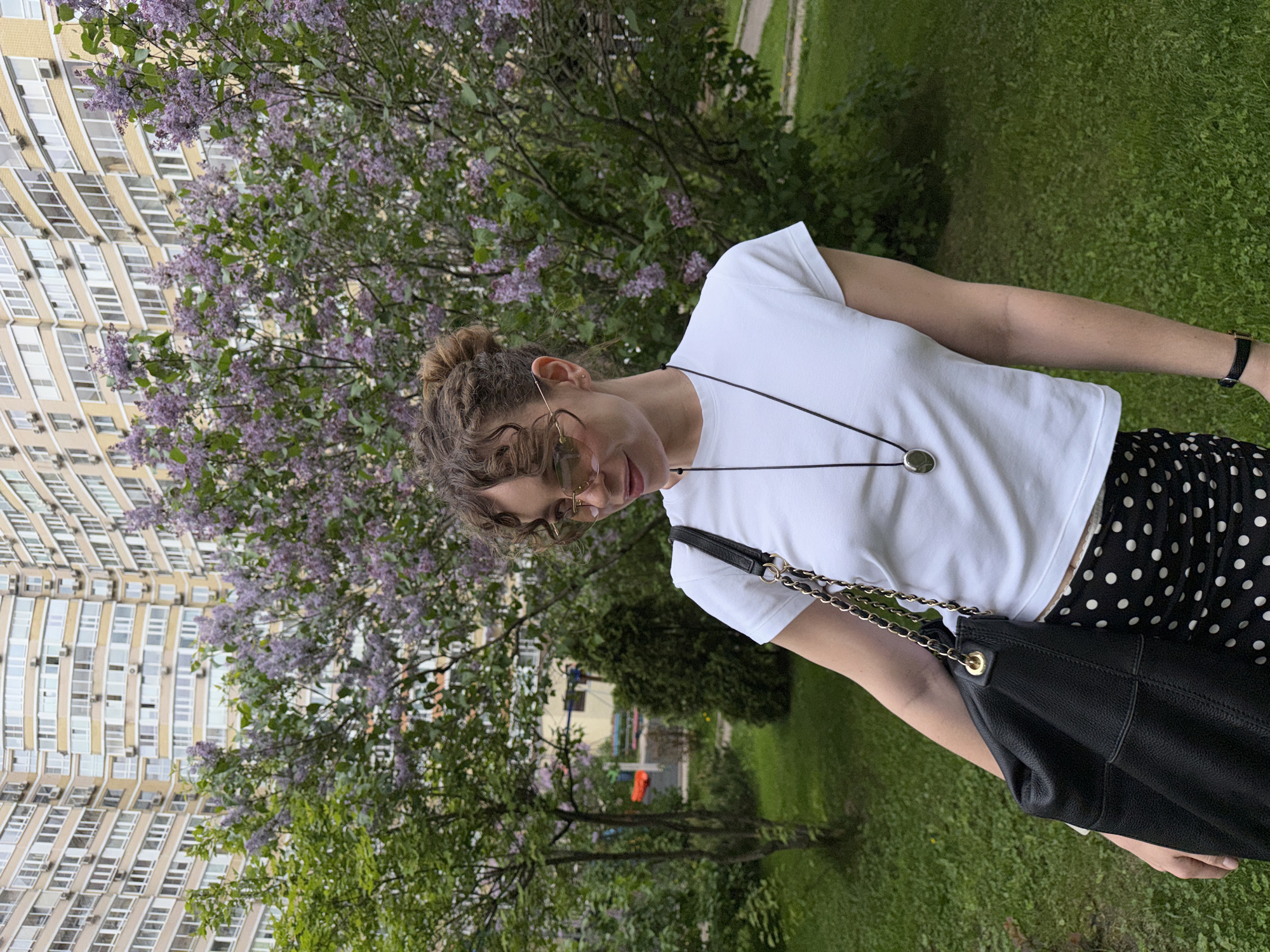 A young adult woman stands outdoors in Moscow, Russia. She wears a white t-shirt, black and white polka dot pants, and glasses. A black purse hangs from her shoulder. A long necklace adorns her neck. The background contains a large building, blooming purple lilac bushes, trees, and green grass.