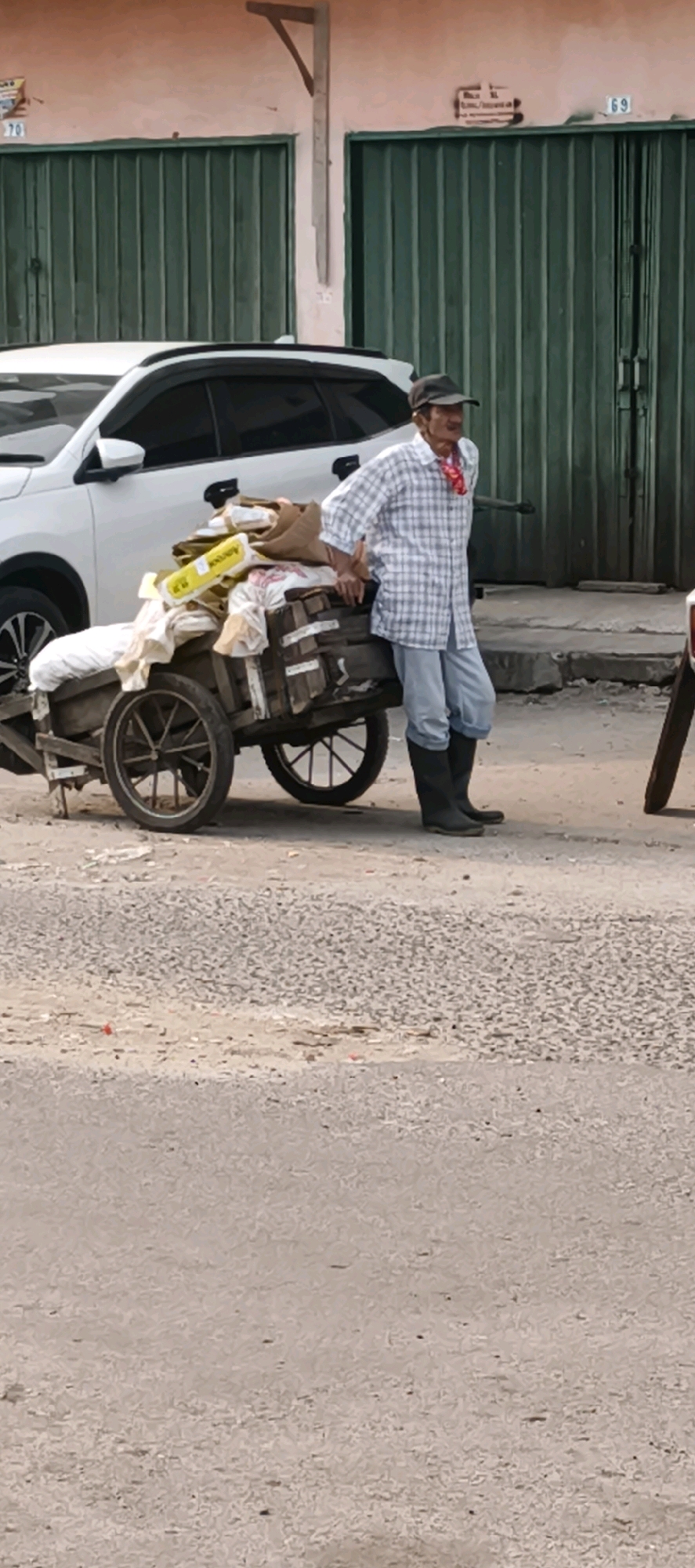 A man stands next to a two-wheeled cart laden with goods, resting his left arm on the cart. He is wearing a plaid shirt, light blue pants, black boots, and a dark baseball cap. A red cloth is tied around his neck. The cart is made of wood and contains various items covered by white fabric and brown bags. Behind the cart and man is a white SUV. The background shows a building with green metal shutters and some signage. The ground is paved with asphalt and gravel. The scene appears to be a street in Jambi, Indonesia.