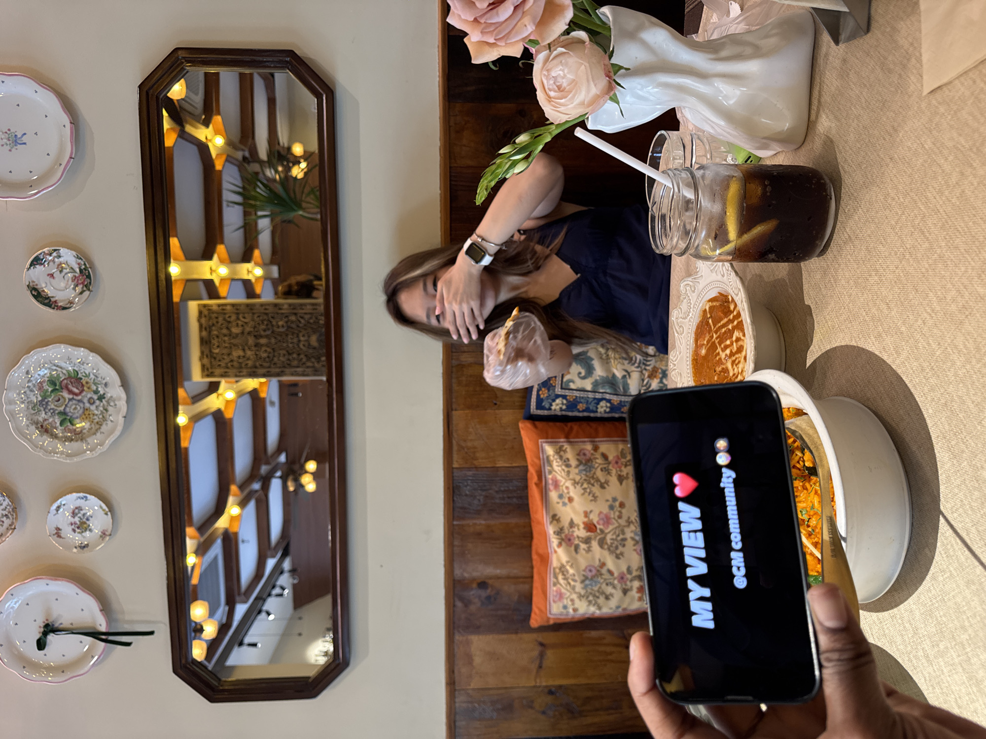 The image shows a table setting with several items. A woman is seated at the table, partially obscuring her face with her hands. She is wearing a watch and a dark-colored top. On the table, there is a vase with peach-colored roses, a glass jar filled with a dark beverage, and two white bowls containing food. One appears to contain a baked dish, and the other holds a dish that looks like fried rice. An individual is holding a smartphone in the foreground, displaying a photo of the food with the text "MY VIEW @CM community" and a heart emoji. A decorative mirror hangs on the wall, reflecting interior architectural details, including lighting fixtures and wooden beams. Decorative plates are also mounted on the wall. The overall setting suggests a restaurant or cafe interior. It is not possible to determine from this photo if this image was taken in the Yuzhong District, China.