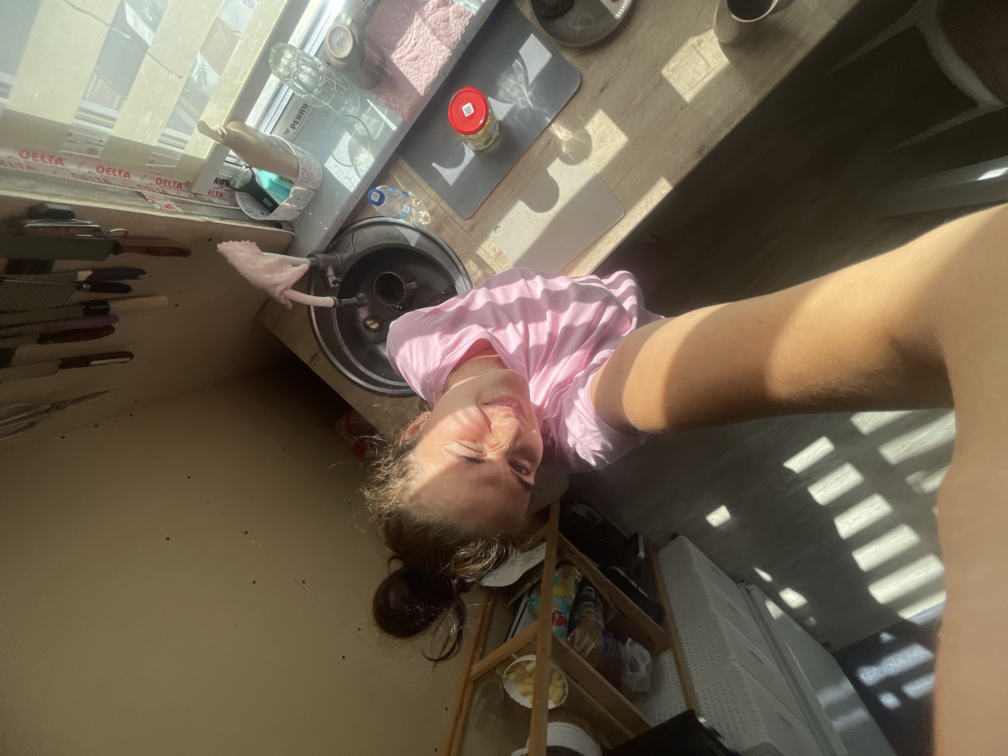 The media file depicts a young woman taking a selfie in a kitchen. She is wearing a pink t-shirt and has her hair pulled back. The woman appears to be smiling and making a playful expression.
The kitchen setting is visible with various objects, including a sink, a wooden countertop, shelves with bottles and bowls, and a window with blinds that cast striped shadows across the scene. There is a knife block with several knives visible on the left side of the image. A jar with a red lid is on a grey mat on the countertop. A water bottle is also on the countertop.
The lighting suggests it is daytime, likely morning or afternoon, with sunlight streaming through the window. The overall mood is casual and lighthearted. The location context provided is "Мосметаллоконструкция Онт, Russia".