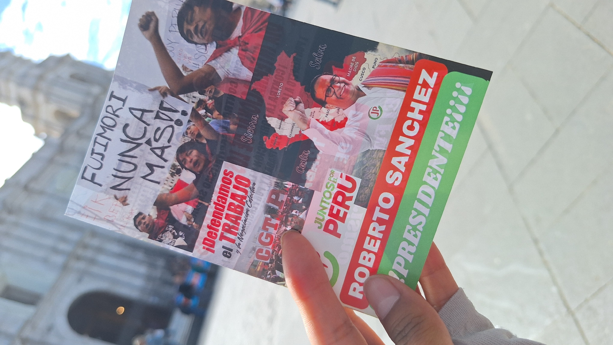 A hand holds a political flyer outdoors, obscuring much of the foreground. In the blurred background, a large, light-colored building with architectural details, possibly a cathedral or government structure, is visible under bright daylight.
The flyer is primarily a political campaign material for "ROBERTO SANCHEZ PRESIDENTE¡¡¡¡" (Roberto Sanchez President!!!!), affiliated with the party "JUNTOS POR EL PERU" (Together for Peru), indicated by text and a "JP" logo. A smiling man, presumably Roberto Sanchez, is depicted with a raised fist.
Other sections of the flyer include protest imagery and political slogans. One prominent slogan reads "FUJIMORI NUNCA MAS!!" (Fujimori Never Again!!). Another section states "¡Defendamos el TRABAJO y la Negociacion Colectiva" (Let's defend WORK and Collective Bargaining), alongside the abbreviation "CGT P", likely referring to a labor union. A stylized map of Peru is shown with various regions labeled, including "Sierra," "Loreto," "Selva," "Costa," "Madre de Dios," "Cusco," "Puno," "Piura," "Amazonas," "Cajamarca," "San Martin," "Pasco," and "Junin." The flyer combines a candidate's promotion with strong social and political messaging, characteristic of election campaigns in Peru.