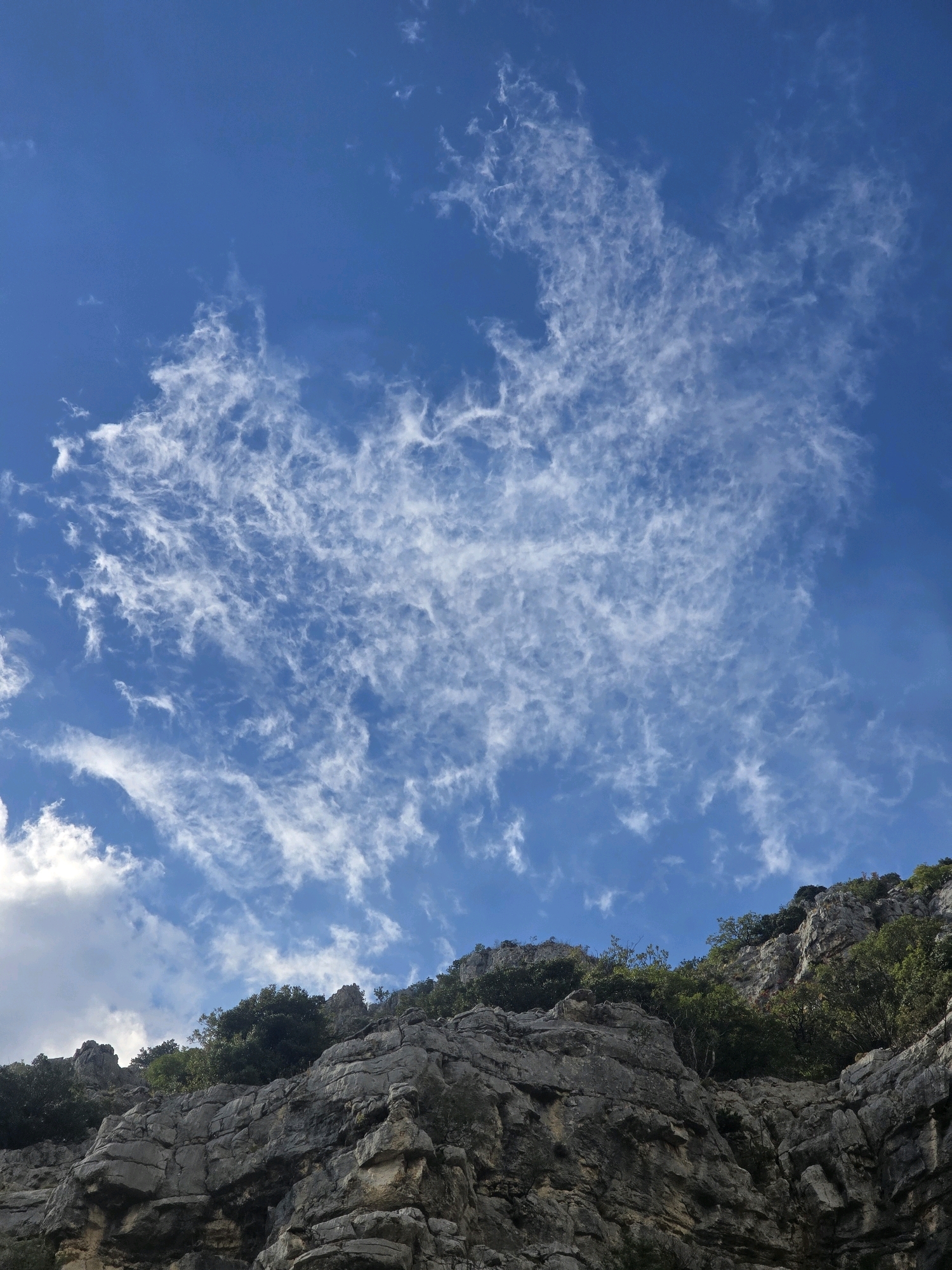 The image displays a low-angle view of a rugged, rocky mountainside with sparse vegetation, set against a bright blue sky filled with wispy white clouds. The rocks appear weathered and layered, with dark green foliage clinging to the slopes. The sky dominates the upper portion of the frame, showcasing a textured cloud pattern. This scene is likely located in a mountainous area surrounding Podgorica, Montenegro, potentially near the Gorica hill or along the edges of the city where natural landscapes meet urban peripheries. There are no people or specific objects visible to indicate human activity within the frame, focusing purely on the natural environment.
