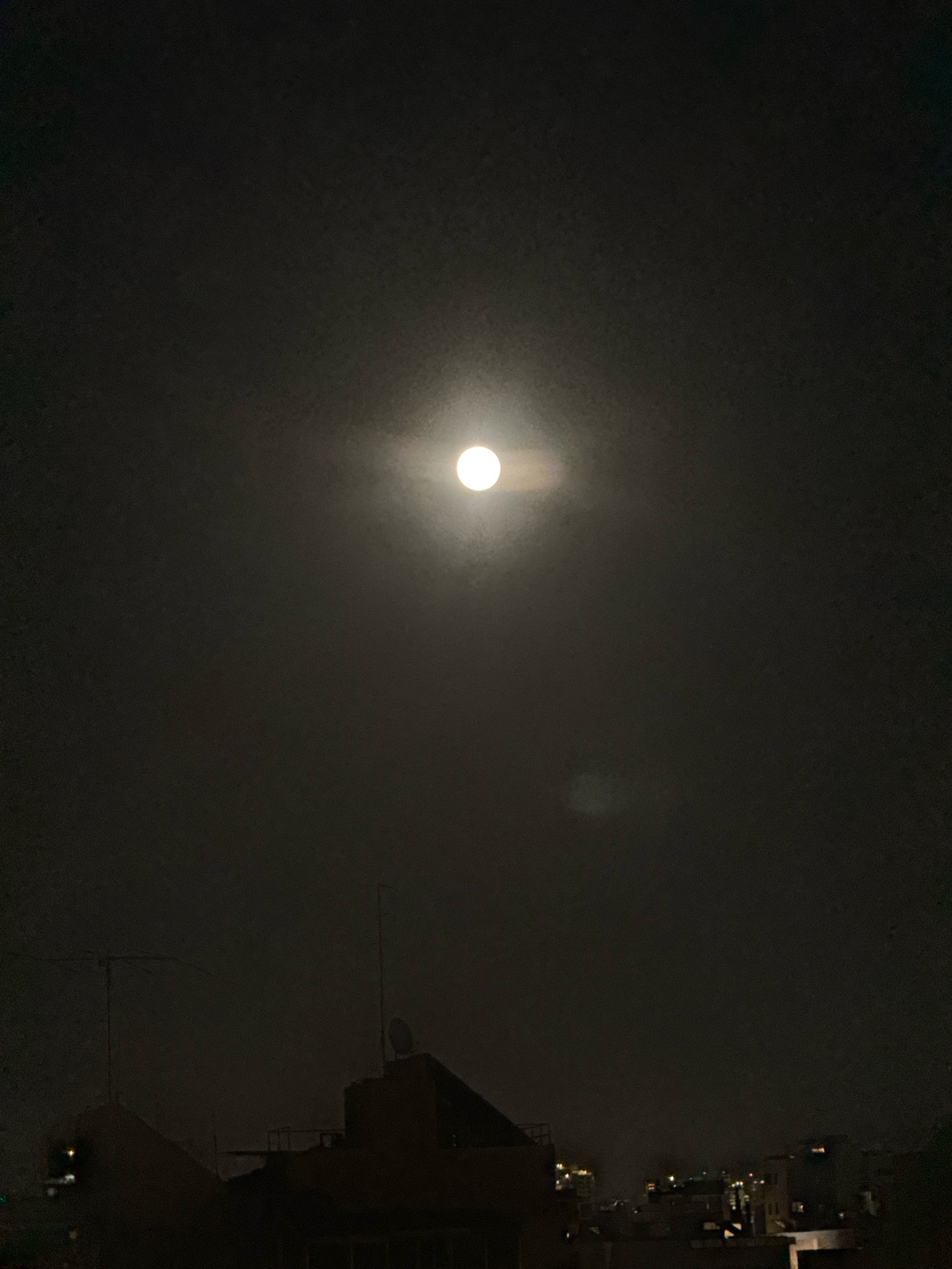 A full moon shines brightly in a hazy, dark sky above Ramat Gan, Israel. The moon is centered in the frame, surrounded by a soft halo of light. Below the moon, silhouetted against the sky, are the rooftops of buildings. Antennas and satellite dishes are visible on some of the rooftops. The overall scene is dark and atmospheric, with the moon being the primary source of light. There are no people or identifiable actions in the image.