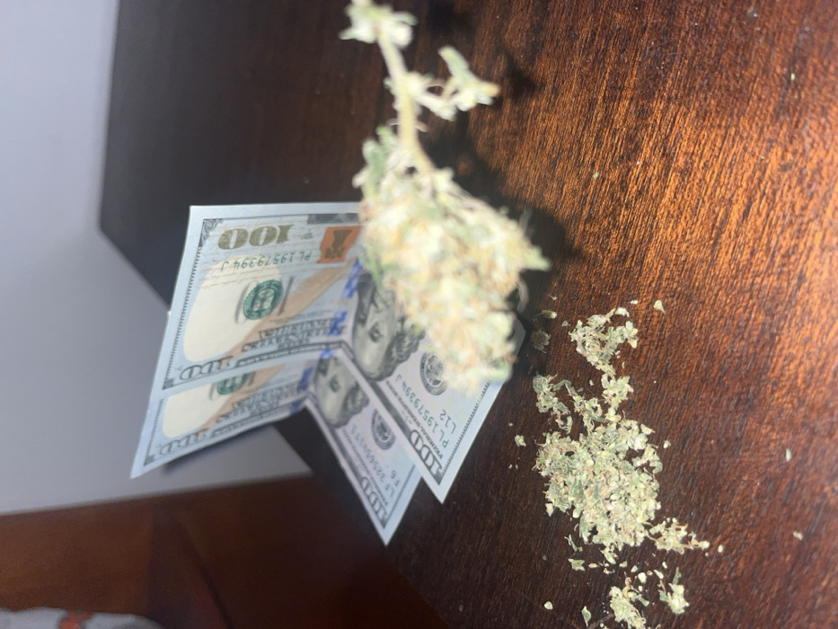 Two hundred-dollar bills are displayed on a wooden surface. A bud of marijuana rests atop the bills, next to some loose marijuana. The scene is somewhat angled and appears to be taken indoors.