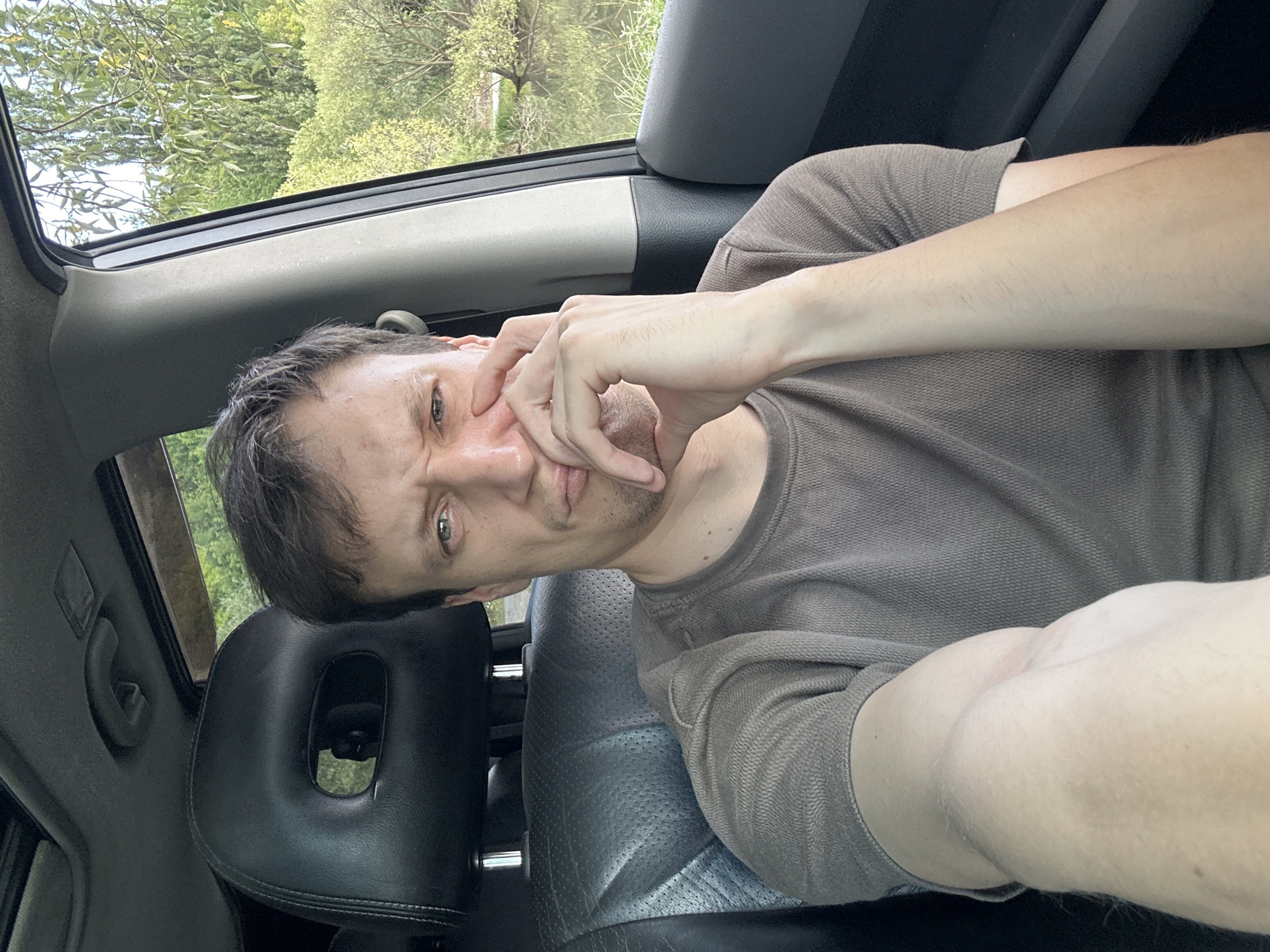 A man is sitting in the driver's seat of a car, facing the camera. He is wearing a brown t-shirt and has his hand on his chin, with a thoughtful or concerned expression. The interior of the car, including the black leather seats and headrest, is visible. Through the open car door and window, a body of water and lush green trees can be seen, suggesting an outdoor, possibly rural or park-like setting. The location is identified as Makiv, Ukraine.