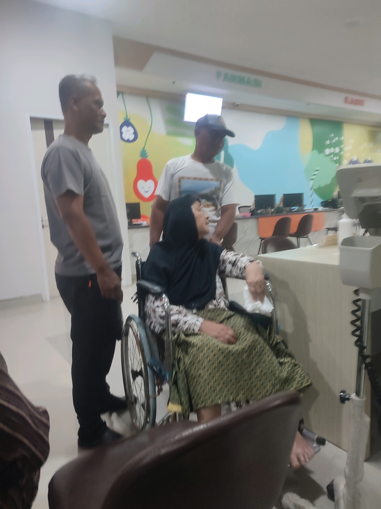 The image depicts an indoor scene, likely a medical facility, given the presence of a wheelchair and a medical equipment stand. A woman wearing a black hijab and a patterned dress is seated in a wheelchair. She is holding a white tissue in her right hand. Two men are present. One man, wearing a grey t-shirt and black pants, stands to the left of the woman. The other man, wearing a white t-shirt and a baseball cap, stands behind and to the right of the woman. In the background, there are desks with computer monitors and chairs, suggesting an office or waiting area. Signs on the wall read "FARMASI" (Pharmacy) and "KASIR" (Cashier), further indicating a retail or service environment within a larger establishment. The overall lighting suggests it might be evening or a dimly lit indoor space. The scene is static, with no overt action occurring beyond the presence of the individuals. The context of Nganjuk, Indonesia, is provided, suggesting the location of this scene.