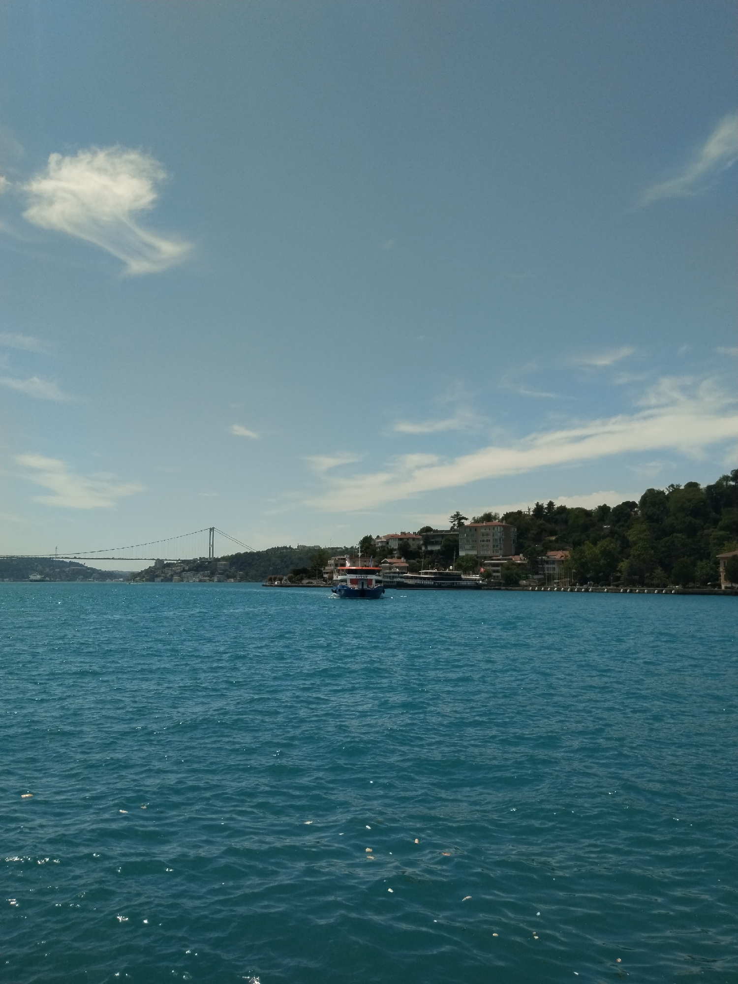 This image captures a wide, scenic view across a vast body of water under a bright, clear blue sky. The water, a vivid deep blue and turquoise, exhibits gentle ripples and small waves across its surface, reflecting the sunlight.
In the middle distance, a white and blue passenger boat, resembling a ferry, is prominently featured, heading from right to left. The name "ZATIN" is clearly visible in white letters on its side.
The distant shoreline on the right is characterized by green, tree-covered hills. Numerous buildings, including multi-story structures and houses with red roofs, are nestled amongst the dense foliage and along the water's edge, suggesting a populated coastal area. Further in the background, a large suspension bridge with white towers and cables spans the water, connecting the landmasses.
The sky, occupying the upper half of the frame, is predominantly clear blue, with scattered white, wispy clouds. A distinctive cloud formation is visible in the upper left quadrant. The bright lighting indicates it is a sunny day, likely late morning or early afternoon. The presence of the suspension bridge, the characteristic blue water, and the hilly, developed shoreline strongly suggest the location is the Bosphorus Strait in Istanbul, Turkey. The scene evokes a sense of calm and is typical of a scenic cruise or view along a major waterway.
