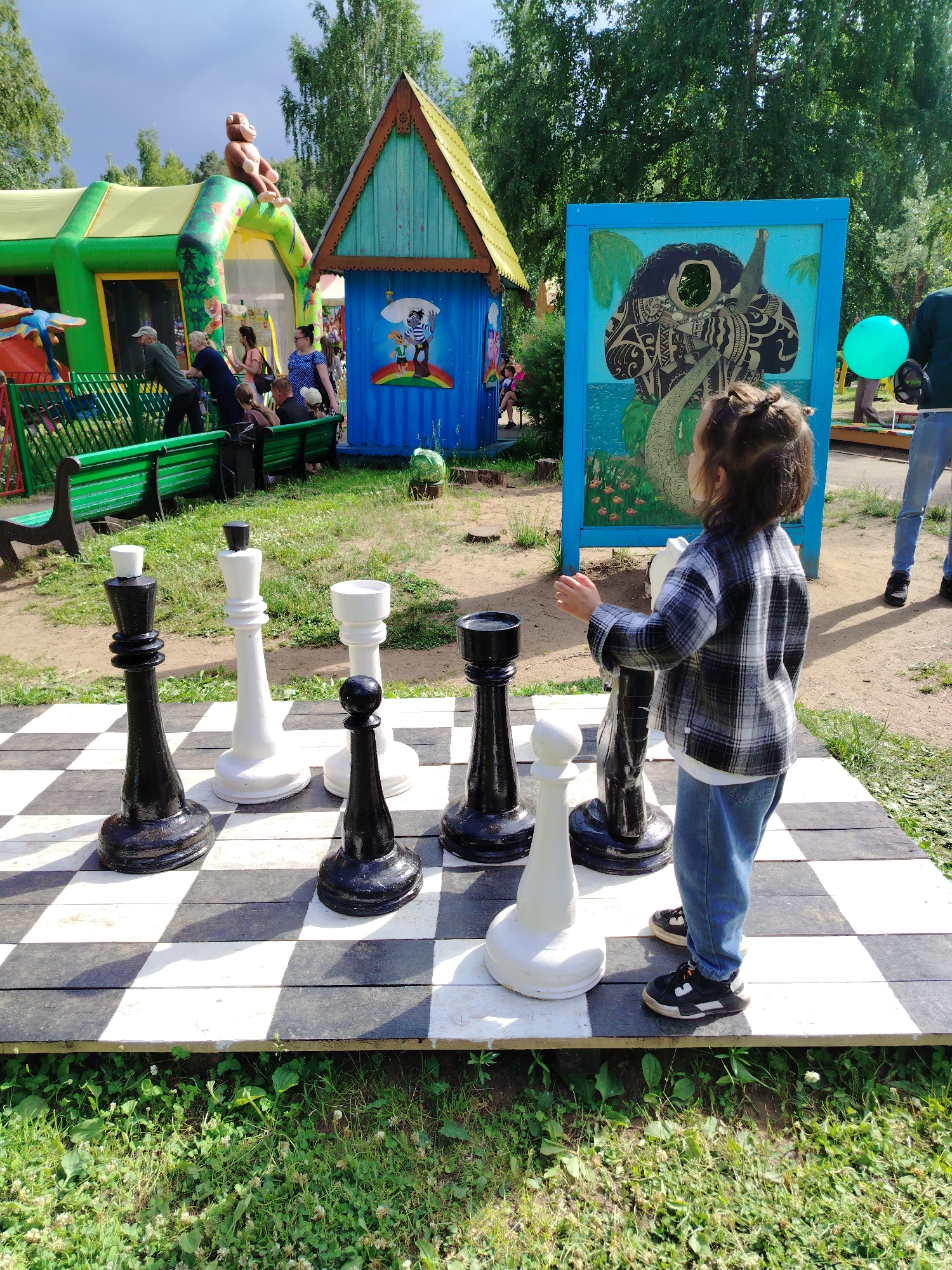 This image depicts an outdoor recreational area, likely a park or playground, in the city of Chaikovsky, Russia. In the foreground, a young girl with dark hair tied in a bun, wearing a plaid shirt and blue jeans, stands on a large wooden chessboard. She is positioned to the right of the chessboard, facing towards a colorful mural on a blue board behind it. The chessboard features oversized black and white chess pieces, including a king, queen, and pawn visible. The background shows a green inflatable play structure with a monkey figure on top, a small blue wooden playhouse decorated with cartoon characters, and a cluster of trees. Several adults and children are visible in the mid-ground, some seated on a green bench near the inflatable structure, while others are interacting in the area. The ground is covered with grass and dirt. The sky is partly cloudy, suggesting it might be a summer day.