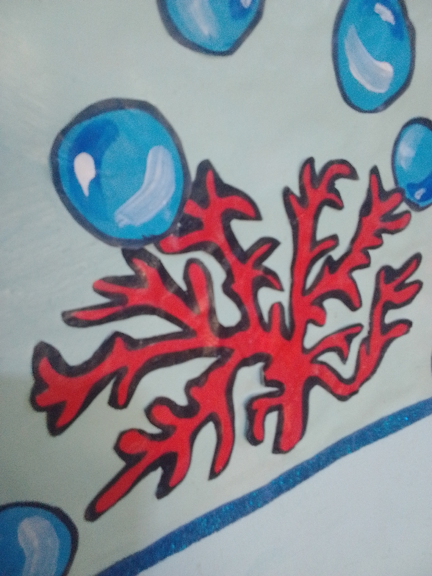 The image displays a drawing of red coral with blue bubbles against a light blue background. The coral is intricate and branching, with a thick black outline. The bubbles are round and glossy, with white highlights, also outlined in black. A glittery blue line runs diagonally across the bottom of the image. The style suggests it could be part of a mural or artwork, possibly for a children's area, given the simplified shapes and vibrant colors. There is no discernible information about the location in La Tagua, Venezuela, as the image only shows the artwork itself.