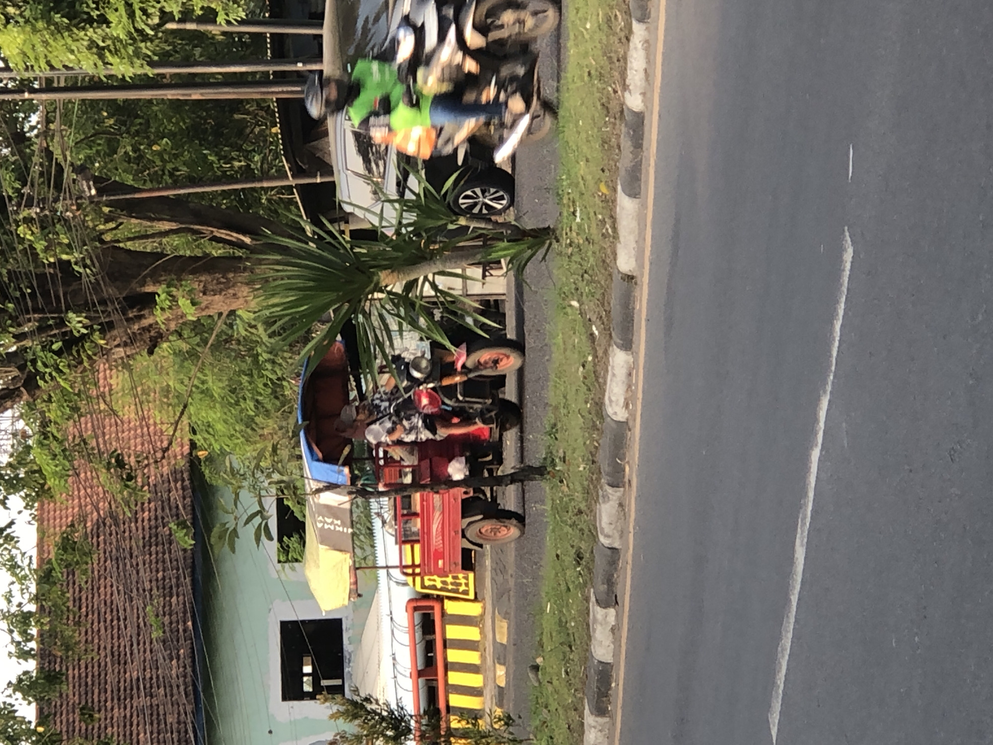 A man rides a red three-wheeled auto rickshaw with a blue canopy, carrying what appears to be a passenger, down a paved road. The auto rickshaw has a small cargo bed behind the driver's seat. To the right of the auto rickshaw, a person in a green vest and helmet rides a motorcycle, also on the paved road. A dark-colored SUV is parked behind the motorcycle. The background shows a building with a tiled roof, lush green trees, and utility poles. The scene is set on a street in Kludan, Indonesia.