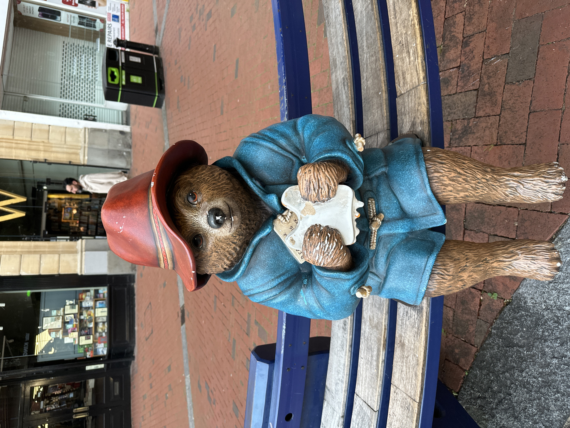 A statue of Paddington Bear, wearing a red hat and a blue coat, sits on a wooden bench in a pedestrian area. The bear is holding a white object in its paws, possibly a map or a book. The background shows shopfronts and a brick-paved walkway. The statue is positioned centrally in the frame.