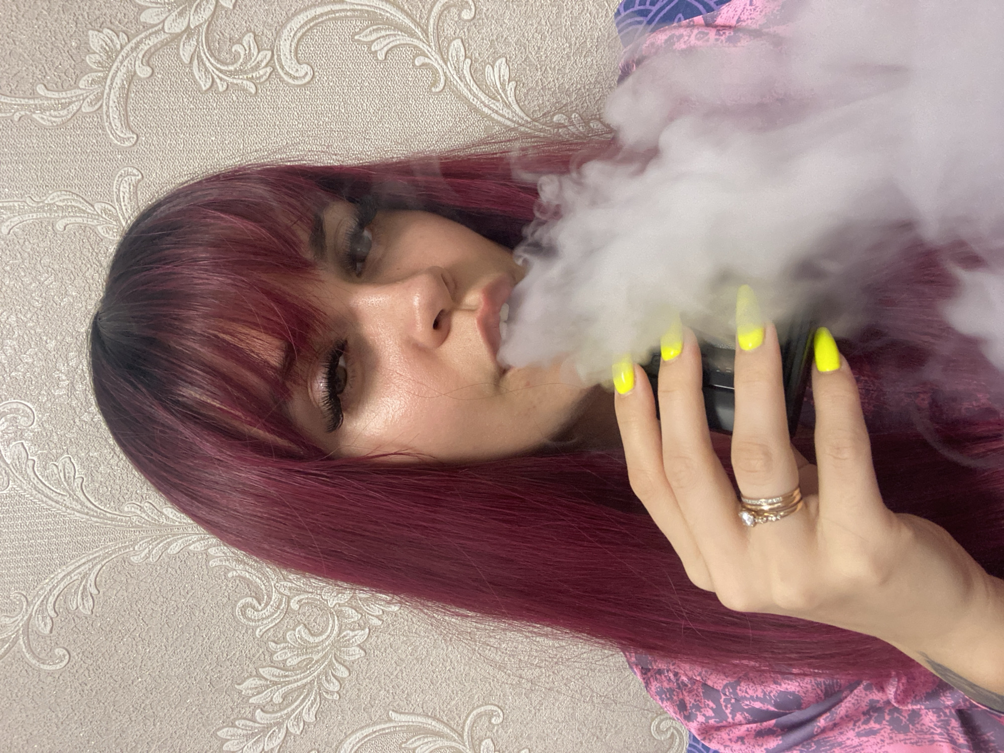 A woman with dark red hair and bangs is lying down, exhaling a large cloud of white smoke. She has neon yellow, long fingernails, and several rings on her finger. She is wearing a pink and blue patterned garment. The background is a light beige wall with a repeating floral pattern. The image does not contain information about the location in the city of Chaikovsky, Russia.