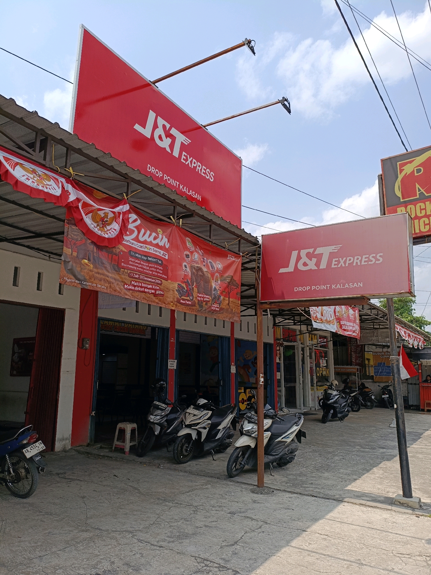 The image depicts a street-level view of a J&T Express drop point located in Maguwoharjo, Indonesia. The primary focus is on the storefront, characterized by a large red J&T Express sign. A smaller J&T Express sign is visible on the right side. Red and white banners hang above the storefront, featuring the Indonesian coat of arms. Several motorcycles are parked in front of the establishment, suggesting it is a busy location. The environment appears to be a commercial area with other businesses visible in the background, including one with an "OPTİK" sign. The sky is partly cloudy, indicating daytime. The overall scene is a typical street view of a logistics service point in an Indonesian urban or suburban setting.