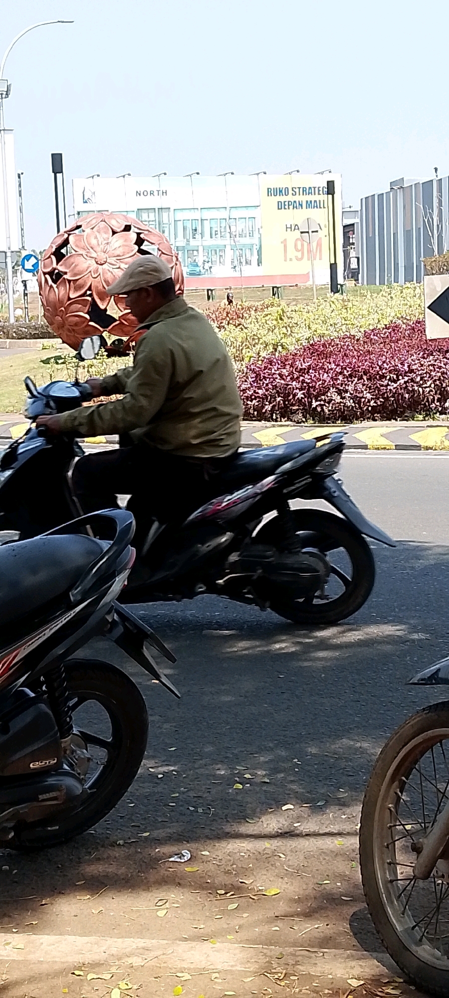 A man wearing a light green jacket and a beige cap rides a black motorcycle on a paved road. The motorcycle is in motion, with the rear wheel visible and blurred. In the background, there is a large, decorative, bronze-colored sculpture resembling a flower. Behind the sculpture, a billboard displays text in Indonesian, including "RUKO STRATEGIC DEPAN MALL" and "HA 1.9M". The billboard also shows an image of a building. To the right of the billboard, a modern building with a striped facade is visible. The ground around the road is covered with sparse green and reddish-purple foliage. The sky is clear and pale. The scene appears to be outdoors in an urban or suburban setting.