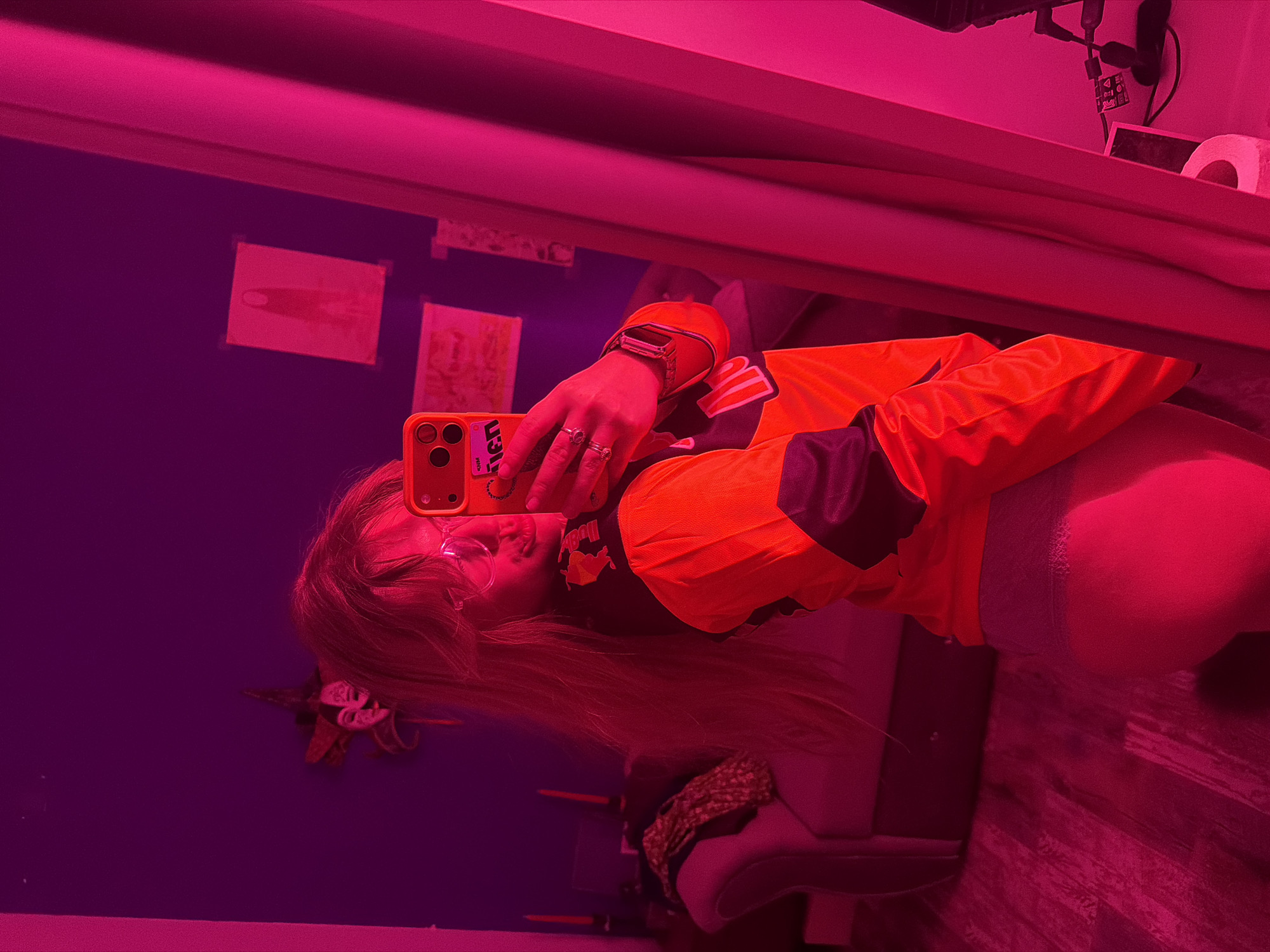 A person is taking a mirror selfie indoors under pinkish-red lighting. The individual has long blonde hair and is wearing glasses and an orange and black long-sleeved garment with some sort of logo. They are holding a cell phone in front of their face, also with an orange case. There are rings on their fingers and a watch on their wrist. The room has blueish-purple walls with pictures taped to them. Behind the individual, a white object, possibly a toilet paper roll, and various electronic cords are visible. A patterned shoe or footstool is visible on the floor. The location appears to be a domestic interior, specifically within the city of Большая Шильна, Russia.