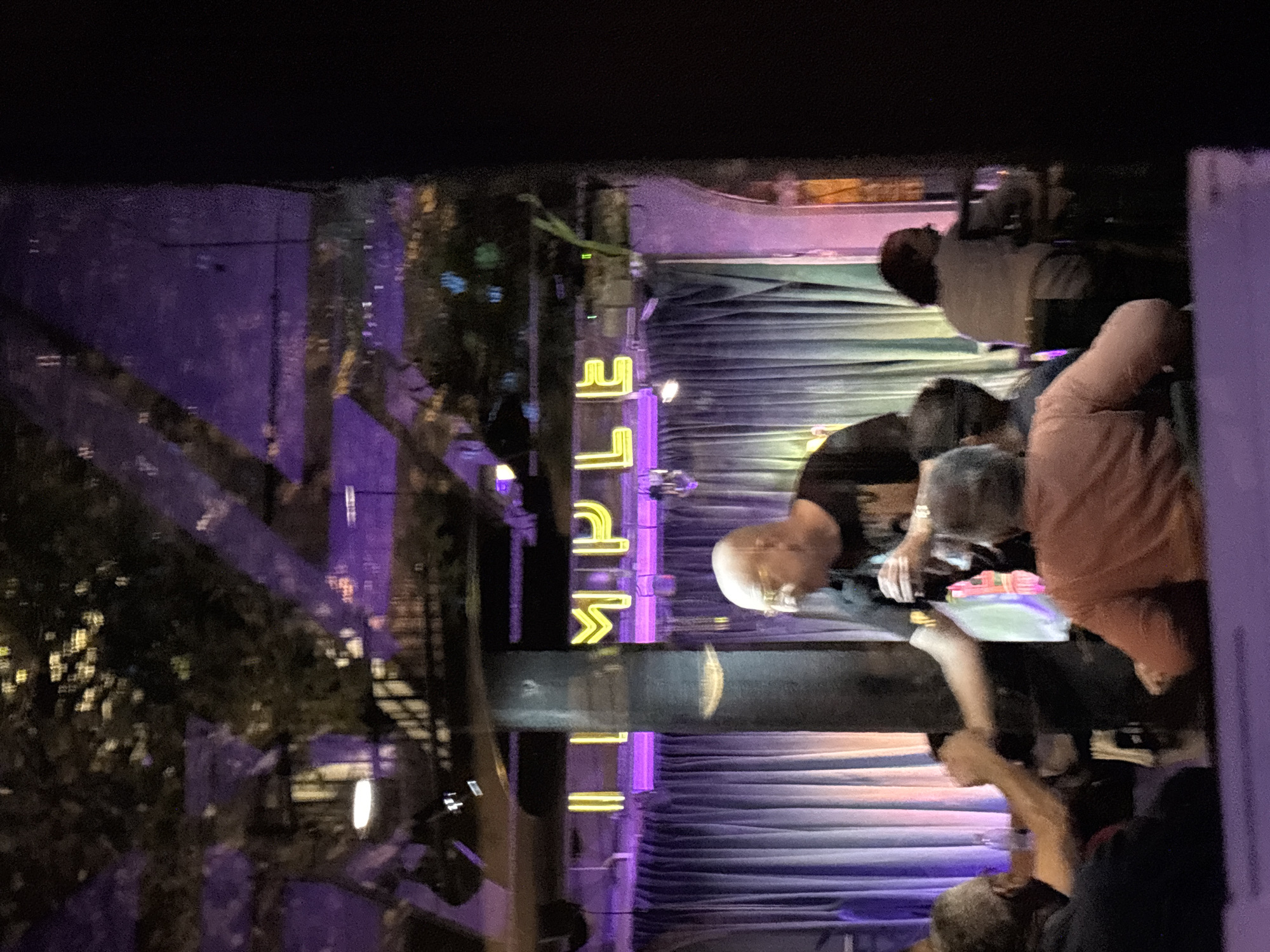 The image depicts a nighttime scene inside what appears to be a bar, club, or performance venue, possibly viewed through a window or reflective surface, given the faint reflections on the left. The overall ambiance is dark, illuminated primarily by artificial lights, particularly vibrant purple and yellow hues.
In the foreground and midground, several individuals are present. A prominent figure is a man with short, light-colored hair and glasses, wearing a dark top, seated or leaning forward, intently looking down at an object or surface in front of him. To his right, another person wearing a reddish-orange or peach-colored shirt is partially visible. Other indistinct figures can be seen further back.
A notable feature of the setting is a bright yellow neon sign in the background, clearly spelling out the word "LIFE". Behind this sign and the people, draped or pleated material, possibly a stage curtain or backdrop, is lit with a strong purple glow. A dark, horizontal barrier or counter runs across the midground, separating the prominent figures from the background elements. The scene conveys a casual gathering or an audience at an event.
The context provided indicates the location is Buenos Aires, Argentina.