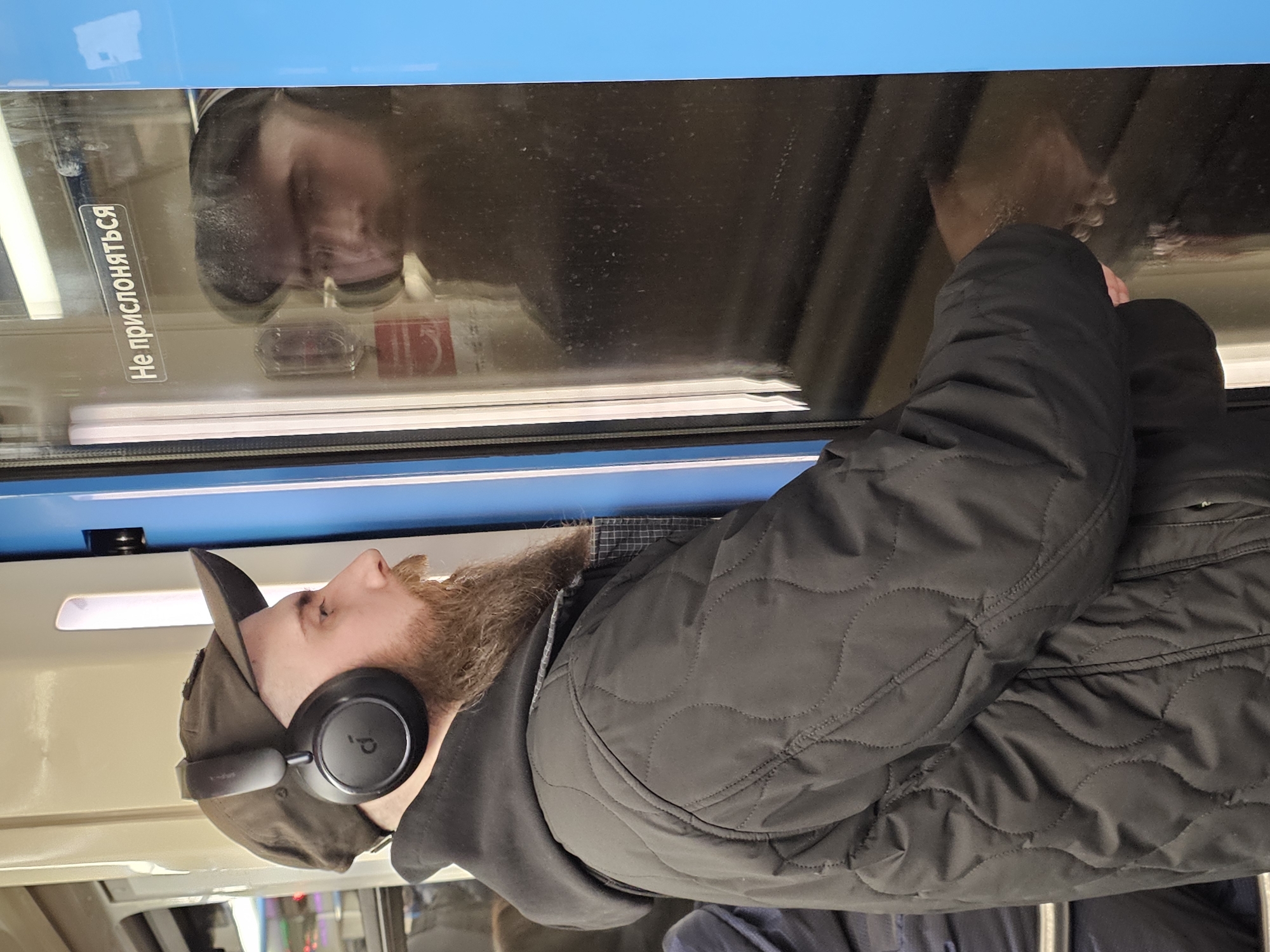 A man with a reddish-brown beard, wearing a dark baseball cap, black over-ear headphones, and a dark quilted jacket, is depicted inside what appears to be a public transport vehicle, likely a train or subway car. He is shown in profile, facing left (after correcting for rotation), seemingly relaxed and possibly listening to music or an audio program.
The man's reflection, along with other indistinct reflections and glare, is visible in the window beside him. The interior of the vehicle features a distinctive bright blue trim above the window and a horizontal white light fixture or trim below it. The jacket has a discernible wavy quilted pattern.
A notable detail is the white rectangular sticker adhered to the window glass, displaying Cyrillic text: "Не прислоняться". This translates to "Do not lean against" or "Do not lean," a common sign found in Russian public transport systems, indicating the location is likely in a Russian-speaking region. The lighting appears to be artificial, suggesting an indoor setting within the moving vehicle. The overall impression is one of a passenger commuting.