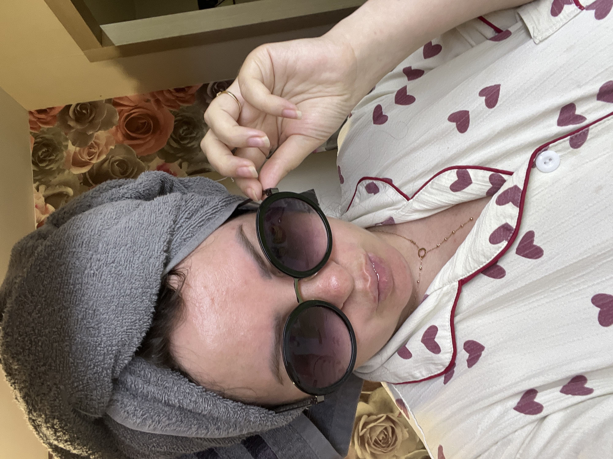 A woman with a grey towel wrapped around her head is wearing sunglasses and pajamas with a heart print. She is holding the sunglasses with her right hand. The background consists of floral wallpaper and a doorway. The location is not specified.