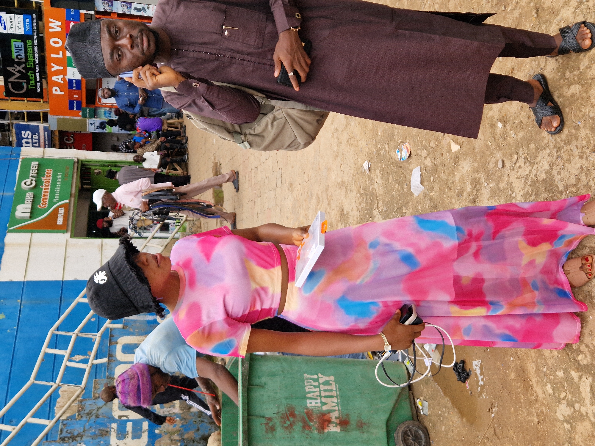 This is a medium shot, outdoors, of a group of people. The image is oriented sideways. A man in the foreground is wearing a dark brown robe and a kufi cap, and he is talking on a cell phone. He has a backpack slung over his shoulder. Behind him, a woman is wearing a black bucket hat and a brightly colored tie-dye dress. She is holding a white object, possibly a tablet or a book. Other people are visible in the background, some sitting and some standing. One person is wearing a light blue shirt and a purple headscarf. A green trash bin with the words "HAPPY FAMILY" is visible in the lower right portion of the image. The ground appears to be dirt or sand. The scene suggests a bustling outdoor area, possibly a market or a busy street, given the presence of commercial signage like "PAYLOW" and "MAHA CITEEM ns Communications". The lighting suggests it is daytime.