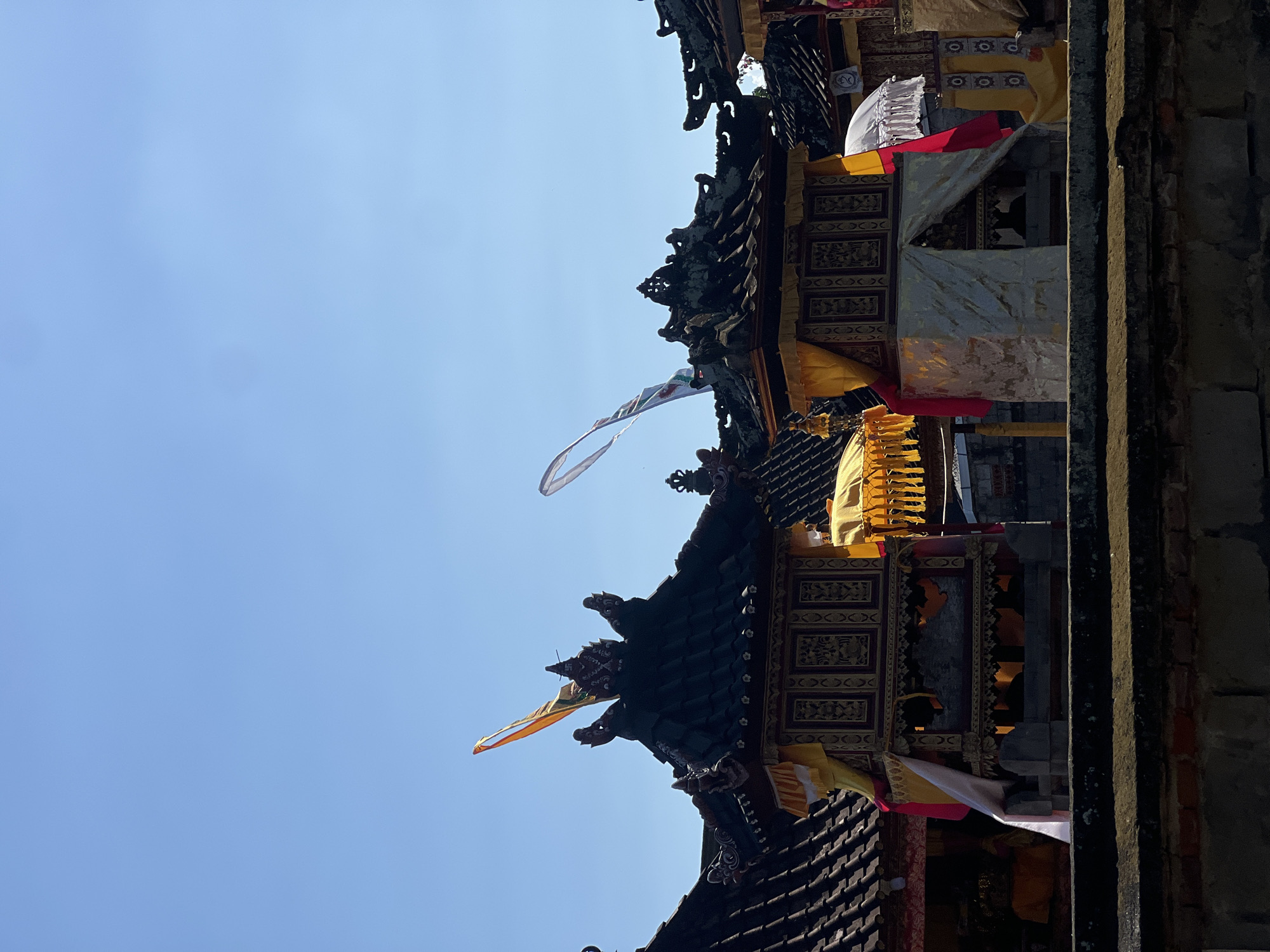 The image shows a street scene in Sukawati, Indonesia, featuring traditional Balinese architecture. Several ornate structures, possibly shrines or parts of a temple, are visible, adorned with colorful fabrics, particularly yellow and white, and decorative banners. The buildings have tiered, dark tiled roofs with intricate carvings. Two flags, one with orange and white elements and another with white and green, are flying against a clear blue sky. The foreground shows a weathered brick wall. The overall impression is of a cultural or religious festival or event taking place.
