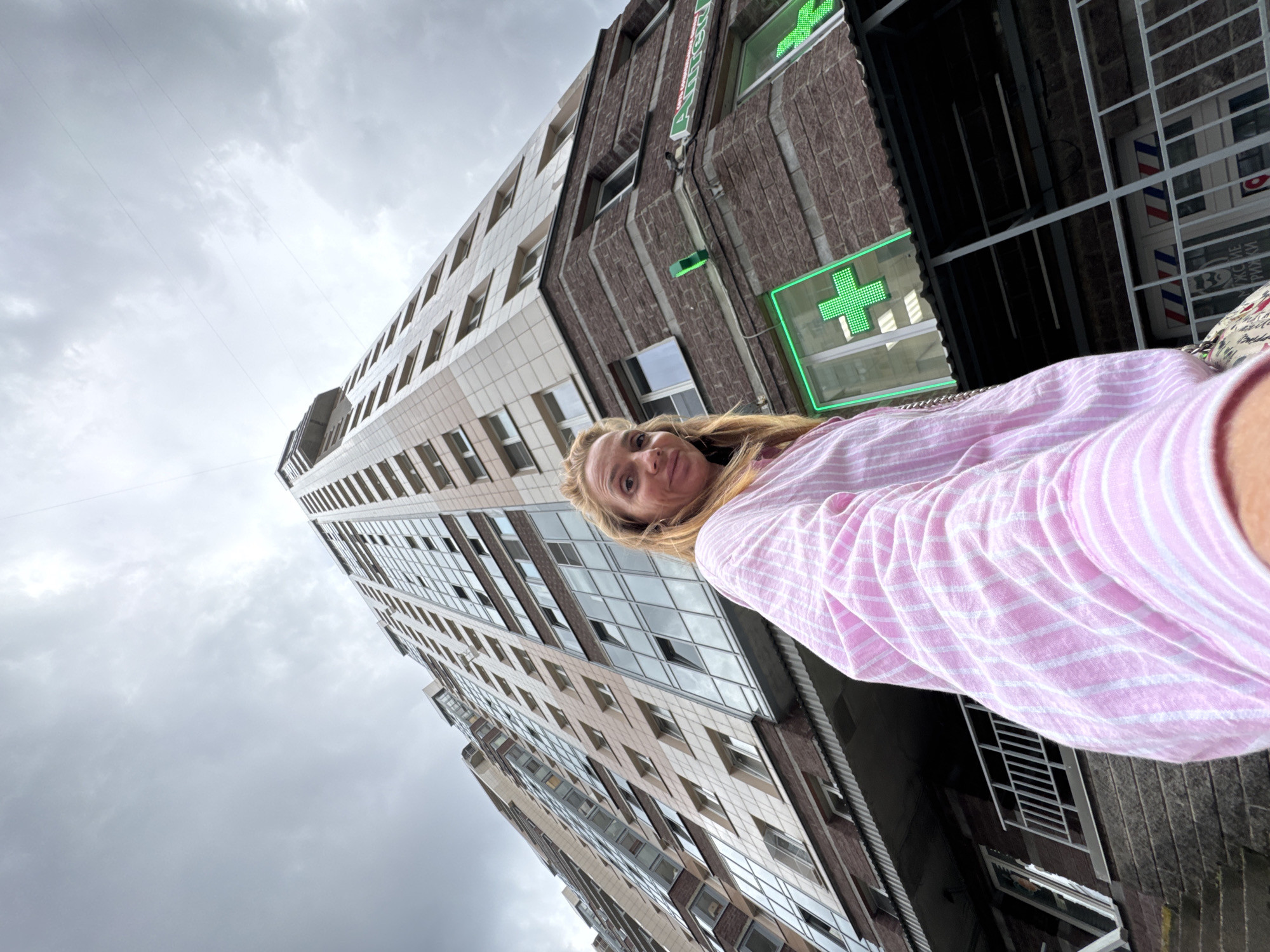 A low-angle, wide-shot selfie captures a blonde woman in a pink and white striped shirt standing in front of a modern, multi-story residential building in Sinyavino, Russia. The building, with its beige and brown brick facade and numerous windows, dominates the background, stretching upwards towards a cloudy, overcast sky. To the right of the building, a pharmacy is identifiable by a bright green cross symbol illuminated on its facade. The woman is positioned on the left side of the frame, facing the camera with a slight smile. Her right arm is extended, holding the camera. The foreground shows a glimpse of a patterned bag or purse on her left side. The overall environment suggests an urban setting with a focus on residential architecture and commercial establishments.