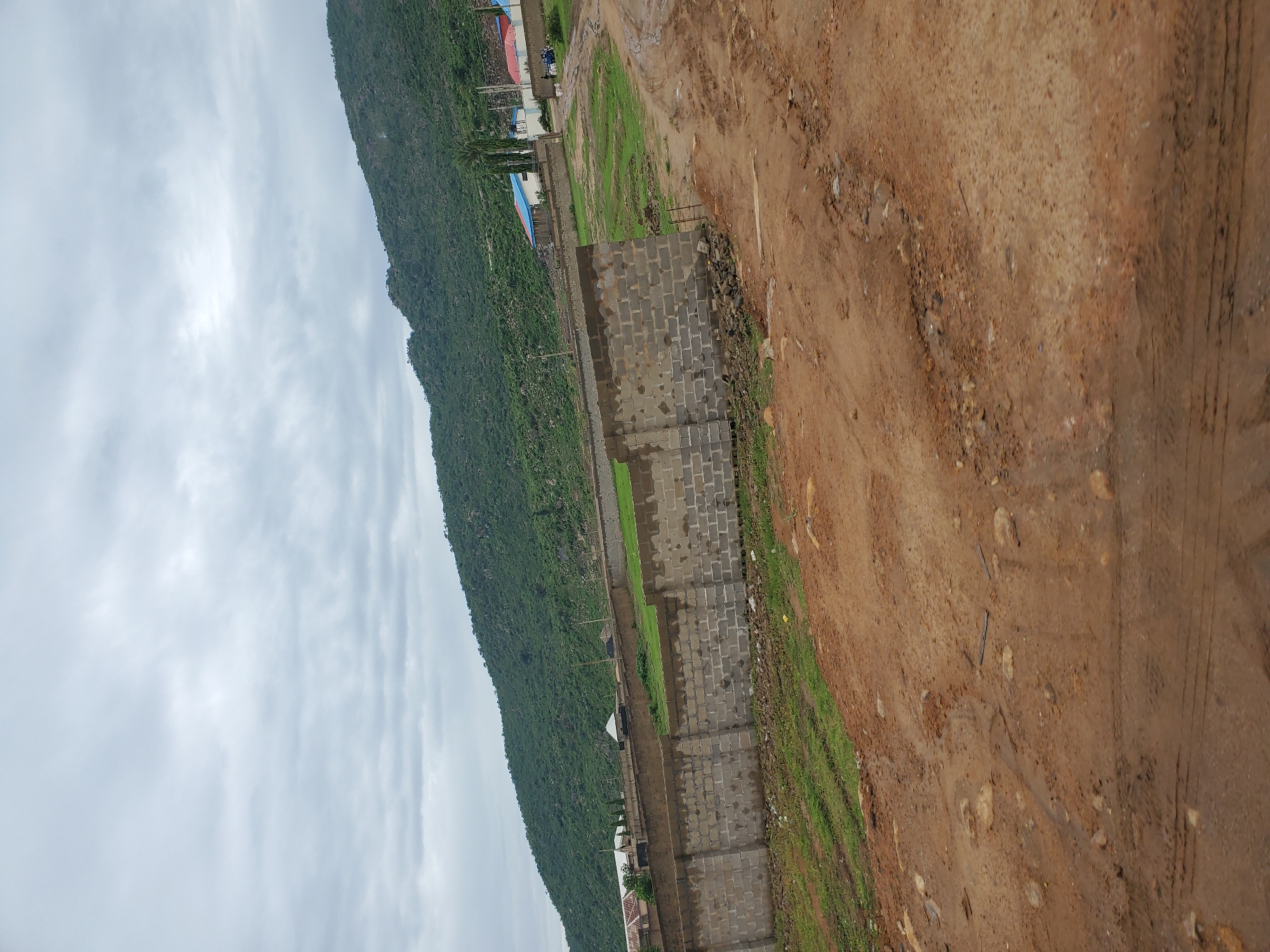 The image depicts a construction site in Naraguta Hausawa, Nigeria, under a cloudy sky. In the foreground, a dirt road with tire tracks and scattered rocks leads into the scene. A partially constructed brick wall runs along the left side of the road, with green grass and some debris on either side of it. Beyond the wall, there are more areas of grass and some scattered trees. In the midground, several buildings with colorful roofs (red, blue, and white) are visible, suggesting a developing residential or commercial area. Some are simple structures with white walls, while others appear more modern with different colored roofs. A few tall, slender trees stand near these buildings. In the background, a large, verdant, and rocky hill or mountain slopes upwards, covered in dense green vegetation and scattered rocks. The overall impression is one of ongoing development in a somewhat rural or peri-urban setting.