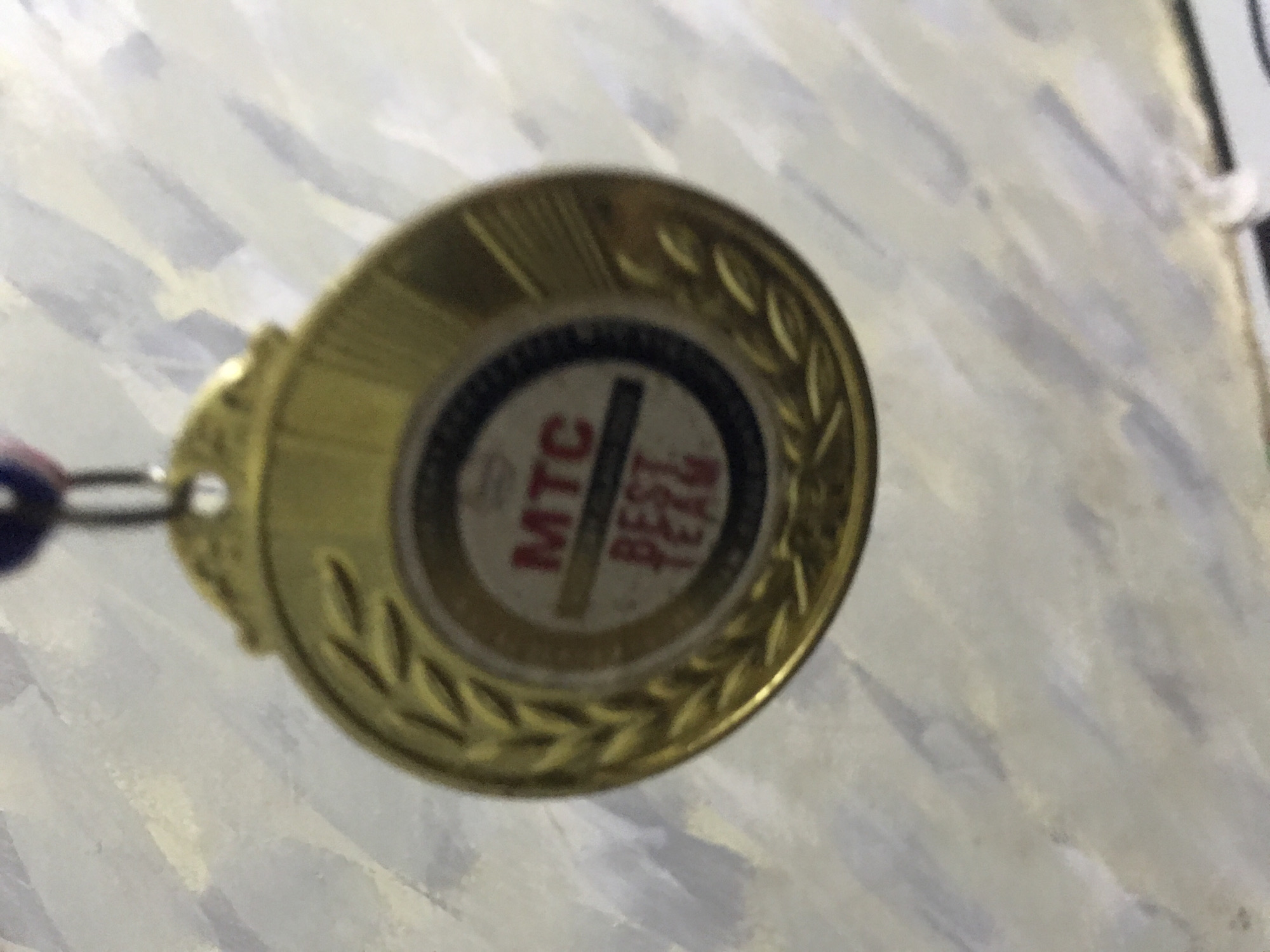 A gold-colored, circular medal is suspended against a textured, light-toned background. The medal features a raised laurel wreath design around its perimeter. The central disc of the medal displays text: "MTC" in red capital letters is positioned above a thin dark horizontal line, and "BEST TEAM" in red capital letters is below the line. This central inscription is set against a light background and framed by a dark ring. A metal jump ring connects the top bail of the medal to a blue and red striped ribbon, part of which is visible at the top center. The background surface exhibits an irregular, brush-stroke-like pattern in shades of light grey, white, and pale yellow. The image is slightly out of focus. The location is Unknown, Unknown.