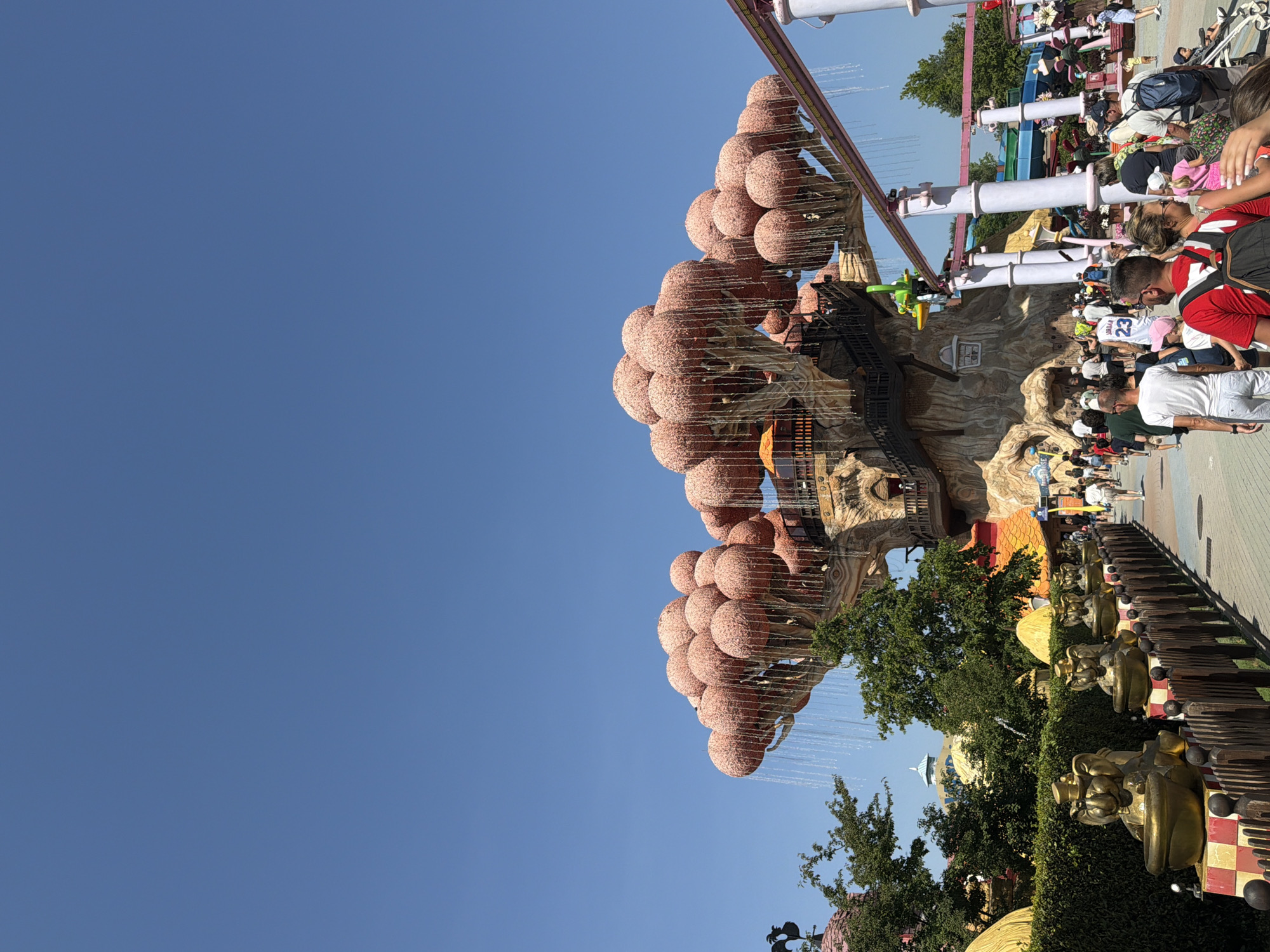 The image depicts a scene at Gardaland, an amusement park located in Castelnuovo del Garda, Italy, under a clear blue sky. The focal point is a large, artificial tree with a trunk resembling bark and a canopy of numerous pink, spherical structures that appear to be stylized fruits or flowers. Water cascades down from these structures, creating a curtain-like effect. A wooden walkway with railings leads towards the tree, and people are walking along it.
In the foreground, a wooden fence separates the walkway from a row of golden statues of cartoonish, elephant-like figures wearing top hats and sitting in bowls. These statues are placed on decorative pedestals. Behind the statues, lush green hedges line the area.
To the right of the tree, a pink roller coaster track is visible, with support pillars. Further in the background, there are various colorful structures and attractions, including what appear to be water slides with blue and green elements. The overall atmosphere is that of a sunny day at a vibrant theme park. The crowd consists of individuals of various ages, dressed in casual summer attire, with some carrying backpacks. The scene captures the lively environment of an amusement park with its distinctive attractions.