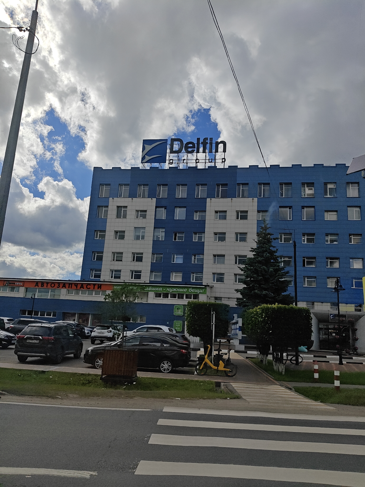 This media file depicts an exterior view of a blue and white multi-story building with a prominent "Delfin Group" logo on top. The building appears to be a commercial establishment, as indicated by the text "АВТОЗАПЧАСТИ" (Auto Parts) and "Клёшка - нужные вещи!" (Klyoshka - necessary things!) visible on lower levels. A street with a crosswalk is in the foreground, with several cars parked and driving. A yellow electric scooter is parked on the sidewalk next to some neatly trimmed bushes. The sky is partly cloudy with patches of blue sky visible, suggesting it is daytime. The overall scene is a typical urban streetscape in Pushkino, Russia, with a focus on commercial activity and public infrastructure.