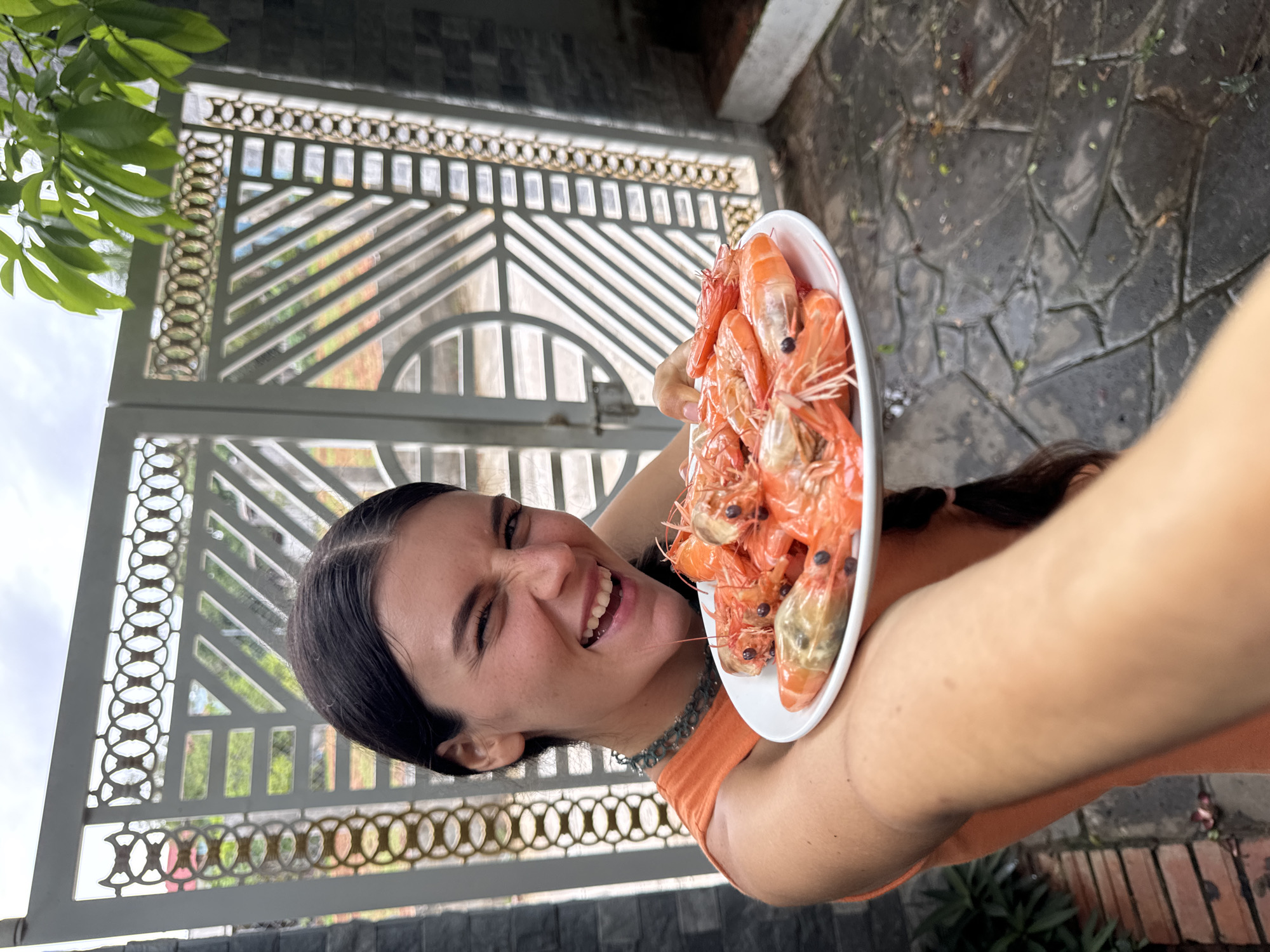 A vibrant, eye-level outdoor shot captures a young woman joyfully presenting a plate of cooked shrimp. The woman, with long dark hair tied back, smiles broadly at the camera, her eyes crinkling with apparent happiness and excitement. She wears an orange sleeveless top and a silver-colored chain necklace.
She holds a white, oval-shaped plate piled high with numerous reddish-orange, cooked shrimp, holding it up towards the viewer, suggesting a selfie perspective. The shrimp appear freshly cooked and appetizing.
The background features a decorative, light-colored metal gate with intricate geometric patterns, including circular and diagonal elements. Through the gate, blurred greenery and distant structures are visible, along with an overcast sky in the upper left corner. The ground in the foreground is paved with textured stone or brick. Green foliage hangs from the top left of the frame, adding to the natural, outdoor setting.
The bright, natural daylight indicates it is daytime. The image conveys a sense of casual enjoyment and anticipation, likely in an outdoor setting in Mũi Né, Vietnam. There is no visible text in the image.