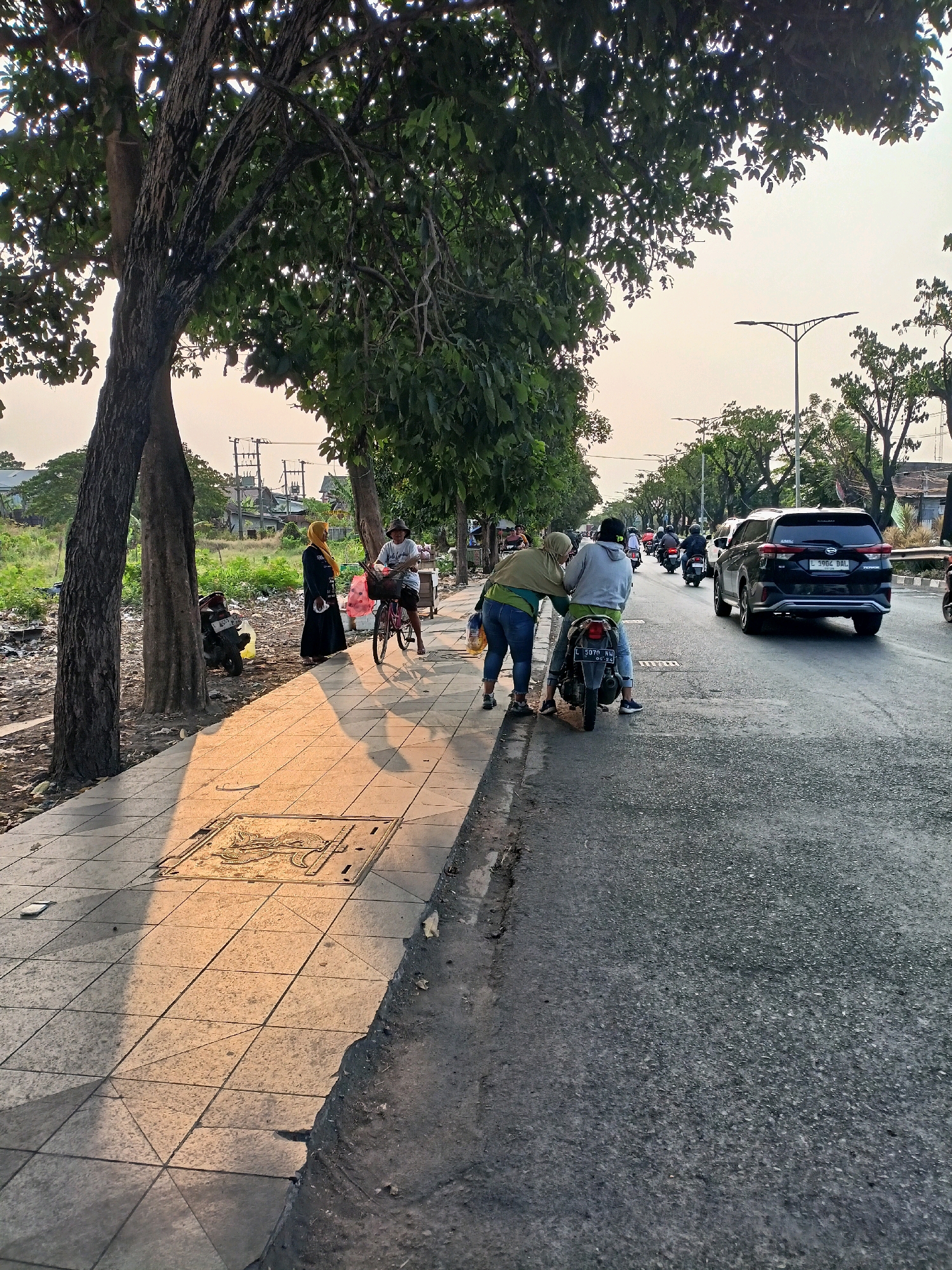 This is a street scene in Surabaya, Indonesia, captured at an angle looking down the road. On the left side of the image is a tiled sidewalk with several trees lining it. In the foreground of the sidewalk, a manhole cover with an embossed design is visible, and the warm glow of late afternoon sun casts long shadows across the pavement. Several people are present: a woman wearing a yellow hijab and a black abaya stands near a parked motorcycle on the sidewalk. Further down the sidewalk, a person on a bicycle with a basket is interacting with another individual. On the right side of the image is a two-lane road with moderate traffic. Several motorcycles and a black SUV are driving on the road. Two individuals are on a motorcycle stopped near the sidewalk, with one person bending over to interact with another person standing beside them. The environment includes greenery, electrical poles, and buildings in the background, indicating an urban setting.