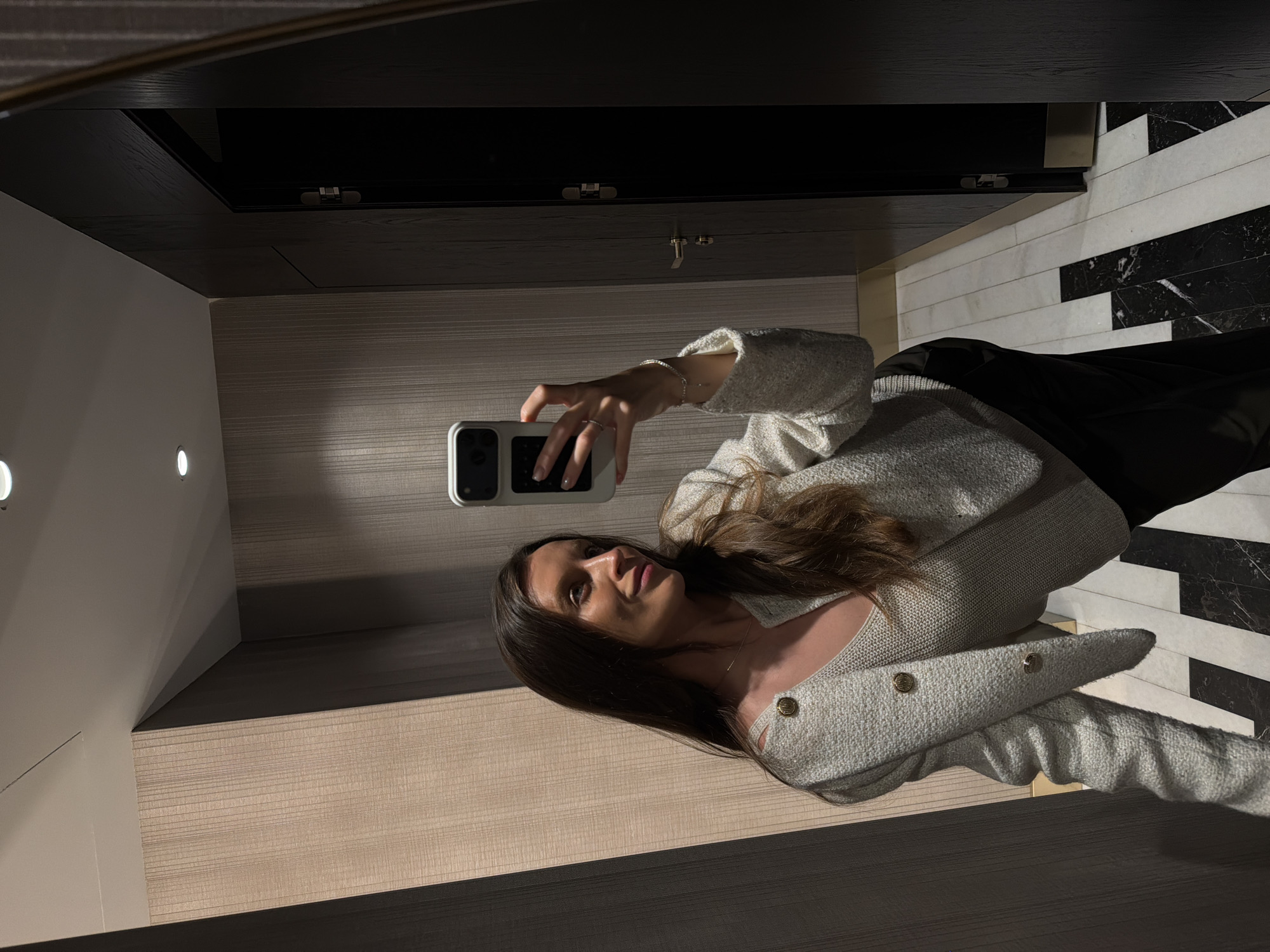 A young woman with long, dark brown hair is captured in a mirror selfie within an upscale interior space in Budva, Montenegro. She is dressed in a light-colored, textured, V-neck cardigan-style jacket adorned with gold-toned buttons, worn over a light ribbed knit top and dark trousers. A thin gold necklace is visible, and she wears a delicate silver-toned bracelet on her left wrist, along with a ring on her left ring finger. Her left hand holds a white smartphone, positioned to capture the reflection. She is looking to her right, away from the camera lens, with a subtle smile.
The environment features light beige, vertically textured wall coverings on the left and front, contrasting with dark brown or black wood-paneled walls and doors on the right. One dark door is slightly ajar, revealing a dark interior and displaying a silver-colored handle and hinge. The ceiling is white with recessed lighting fixtures. The floor consists of a distinctive geometric pattern of alternating black and white rectangular marble tiles.