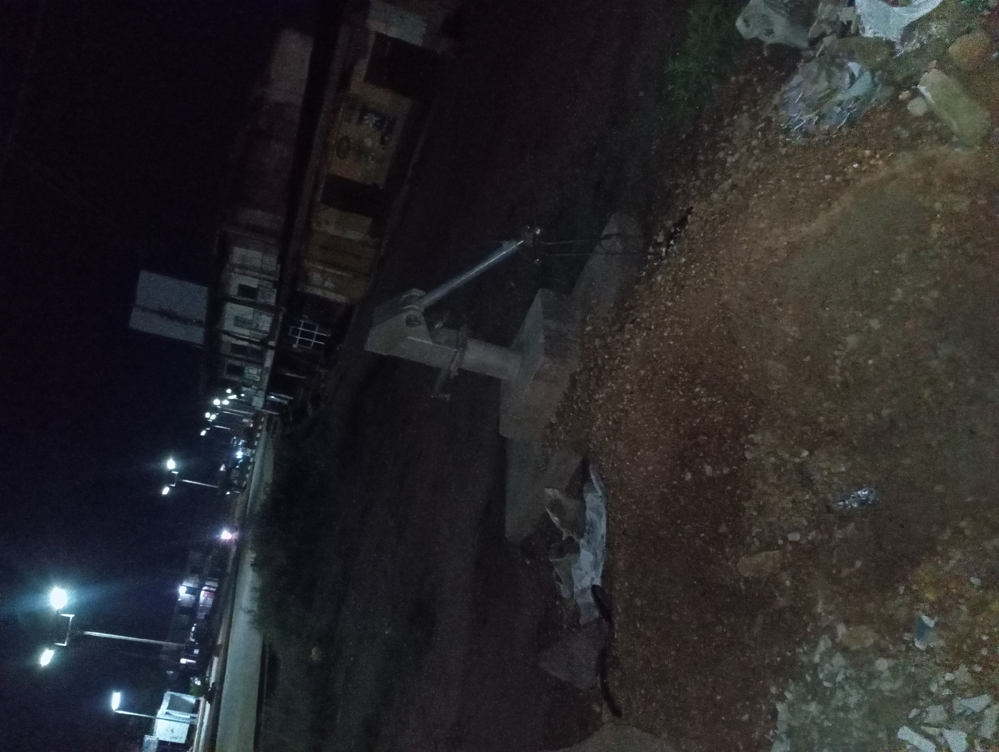In a nighttime urban scene, an old-fashioned hand water pump is situated on a dirt mound. The pump has a grey metal body and a long, slender metal arm, with a pipe extending downwards. The immediate area around the pump is cluttered with debris, including a white tarp, rocks, and some broken white objects. Further in the background, a street is visible, illuminated by streetlights and the lights of buildings. Several structures line the street, some appearing to be shops or residences, with windows and doorways visible. The overall impression is one of a somewhat neglected or undeveloped urban area at night.