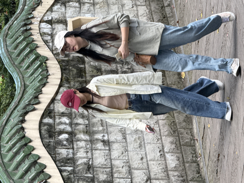 Two females are observed walking on a paved pathway adjacent to a stone wall in Yuzhong District, China. The wall is constructed of grey, irregularly shaped stone blocks, topped with a wavy beige coping and a series of dark green, curved roof tiles. Green foliage is visible above the wall's decorative tiles.
The individual on the left, an East Asian female, wears a maroon baseball cap, a light beige open long-sleeve shirt over a light brown crop top, a dark belt, faded blue wide-leg jeans, and white sneakers. She holds a light-colored smartphone in her right hand and her gaze is directed downwards.
The individual on the right, an East Asian female, wears a light beige baseball cap, eyeglasses, a light grey distressed hooded sweatshirt, faded blue jeans, and light-colored sneakers. She holds a light-colored smartphone in her left hand and has a brown cylindrical cross-body bag slung over her right shoulder. Her gaze is directed slightly upwards and to the right. Scattered dry leaves are present on the pathway.