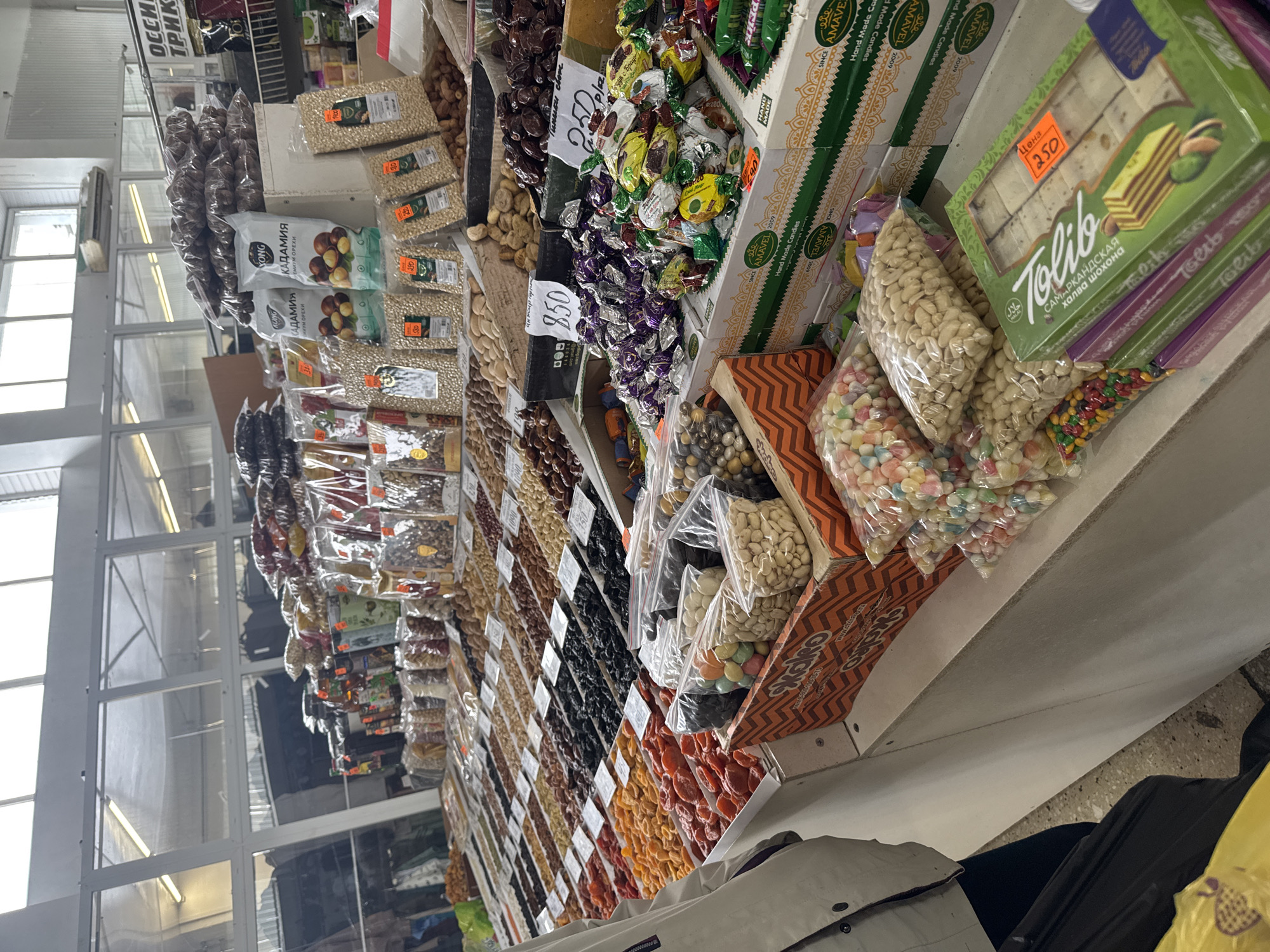 This eye-level, medium shot captures a vibrant market stall, likely located in Chaikovsky, Russia. The stall is brimming with a diverse array of unpackaged and packaged food items, primarily nuts, dried fruits, and confectionery, displayed on multi-tiered shelves.
In the foreground and midground, numerous clear plastic bags contain various types of nuts, including macadamia nuts branded "YONG КАДАМИЯ ПРЕМИУМ ОРЕХИ" (Macadamia Premium Nuts), peanuts, and possibly pistachios. Bags of colorful candies, such as jelly beans and sugar-coated sweets, are also visible. Adjacent to these are boxes of confectionery, prominently featuring "AMAVEL 2009 Hand Made Candies." A stack of green boxes contains "Tolib САМАРКАНДСКАЯ халва шохона" (Tolib Samarkand Halva Shokhona), marked with a "HALAL" logo and a price tag of "Цена 250" (Price 250 rubles). Other orange price tags indicate values like "290" and "850" rubles for various products.
Further down the display, a wide assortment of dried fruits, including bright orange dried apricots and dark prunes/plums, are neatly arranged in rows. Various wrapped candies and chocolates are also packed into boxes. The background reveals large windows with white frames and fluorescent strip lighting, suggesting an indoor market setting with natural light augmenting artificial illumination. Portions of what appear to be concrete flooring and the lower part of a person's leg and belongings (black pants, yellow bag, grey jacket) are visible in the bottom right, hinting at a customer or vendor. The scene depicts a typical commercial activity of selling goods at a local market. Visible Cyrillic text, including "OCCH ТРИК" (partially visible, likely a section name), confirms the Russian-speaking environment.
