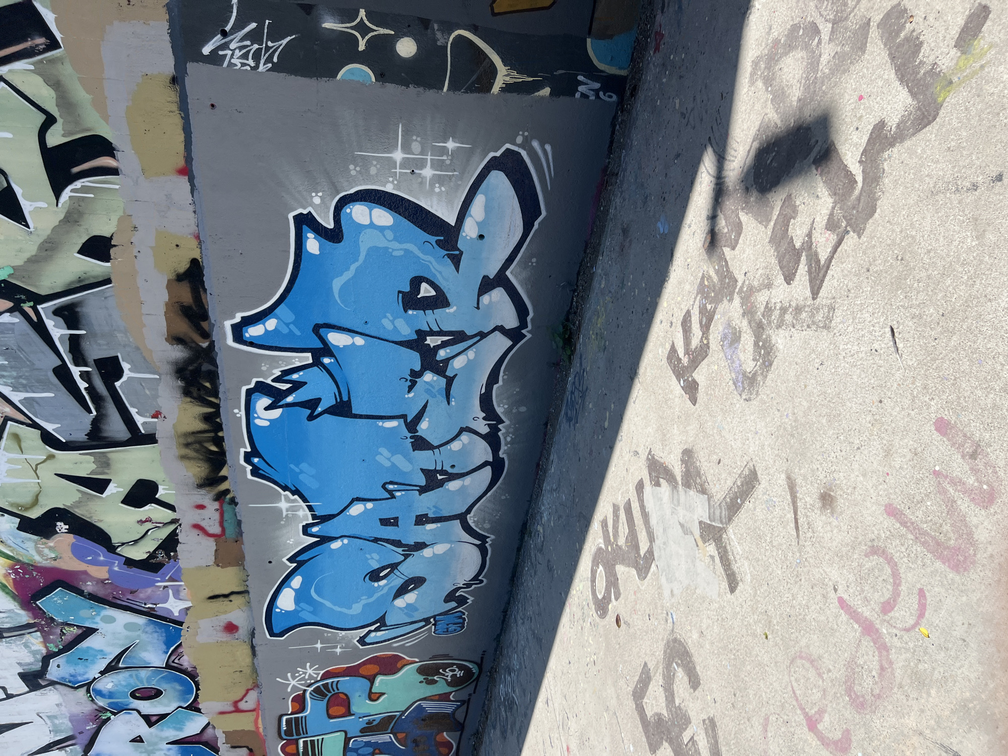 The image depicts a graffiti-covered concrete wall and a section of the ground in Barcelona, Spain. The wall is adorned with vibrant and elaborate graffiti art. In the foreground, the concrete ground is also marked with graffiti and shadows cast by an unseen object. The scene appears to be outdoors, possibly in an urban environment, under bright sunlight.