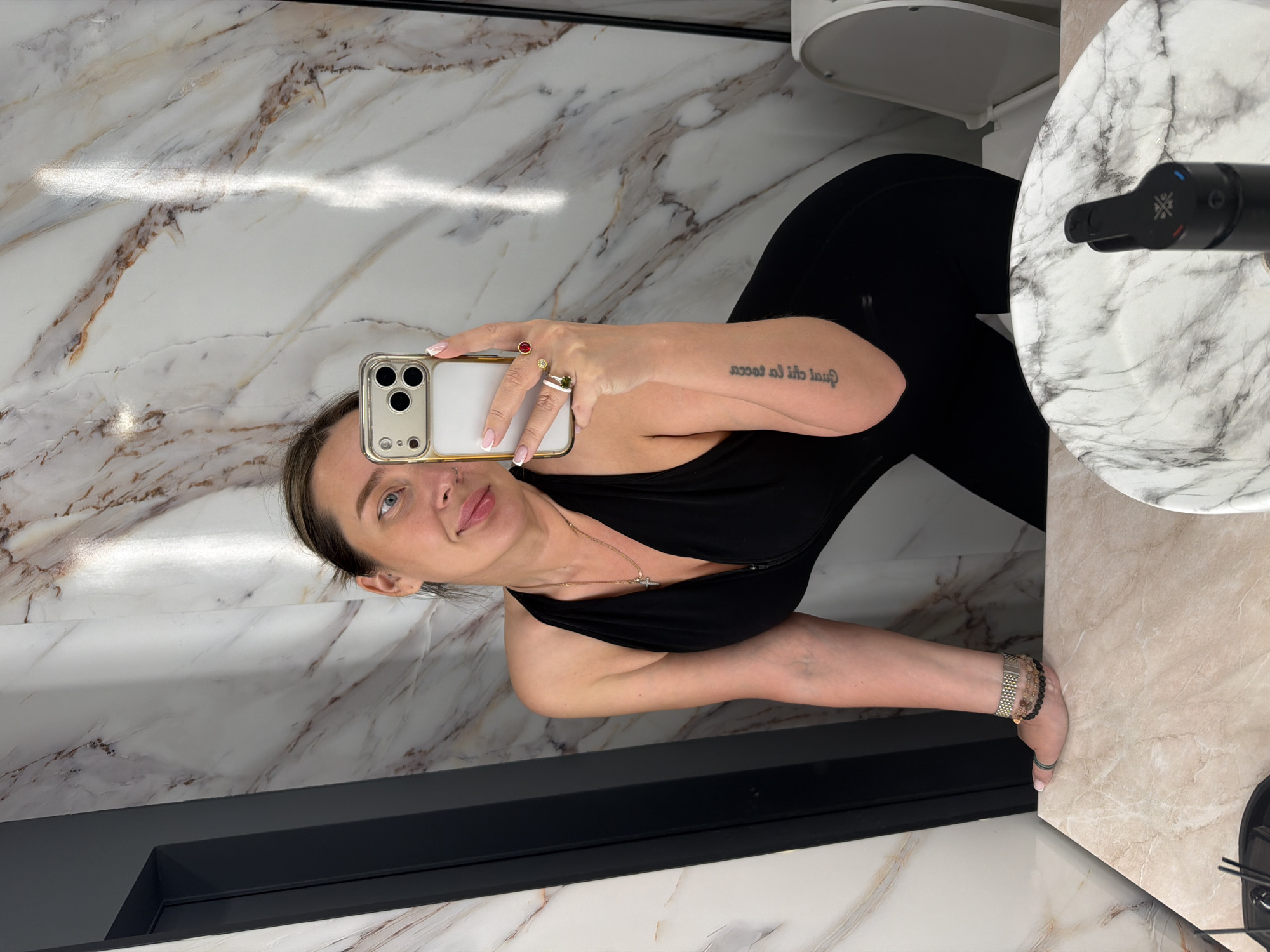 A woman is taking a selfie in a bathroom. She is wearing black leggings and a black top, and holding an iPhone in her left hand, adorned with multiple rings. The woman has a fair complexion, light eyes, and dark hair, and a small tattoo on her left forearm. The bathroom features a marbled wall and floor, a dark grey frame around what appears to be a shower or bathtub area, a white toilet, and a white marble sink with a black faucet. There is no evidence to suggest that the location is in Moscow, Russia.