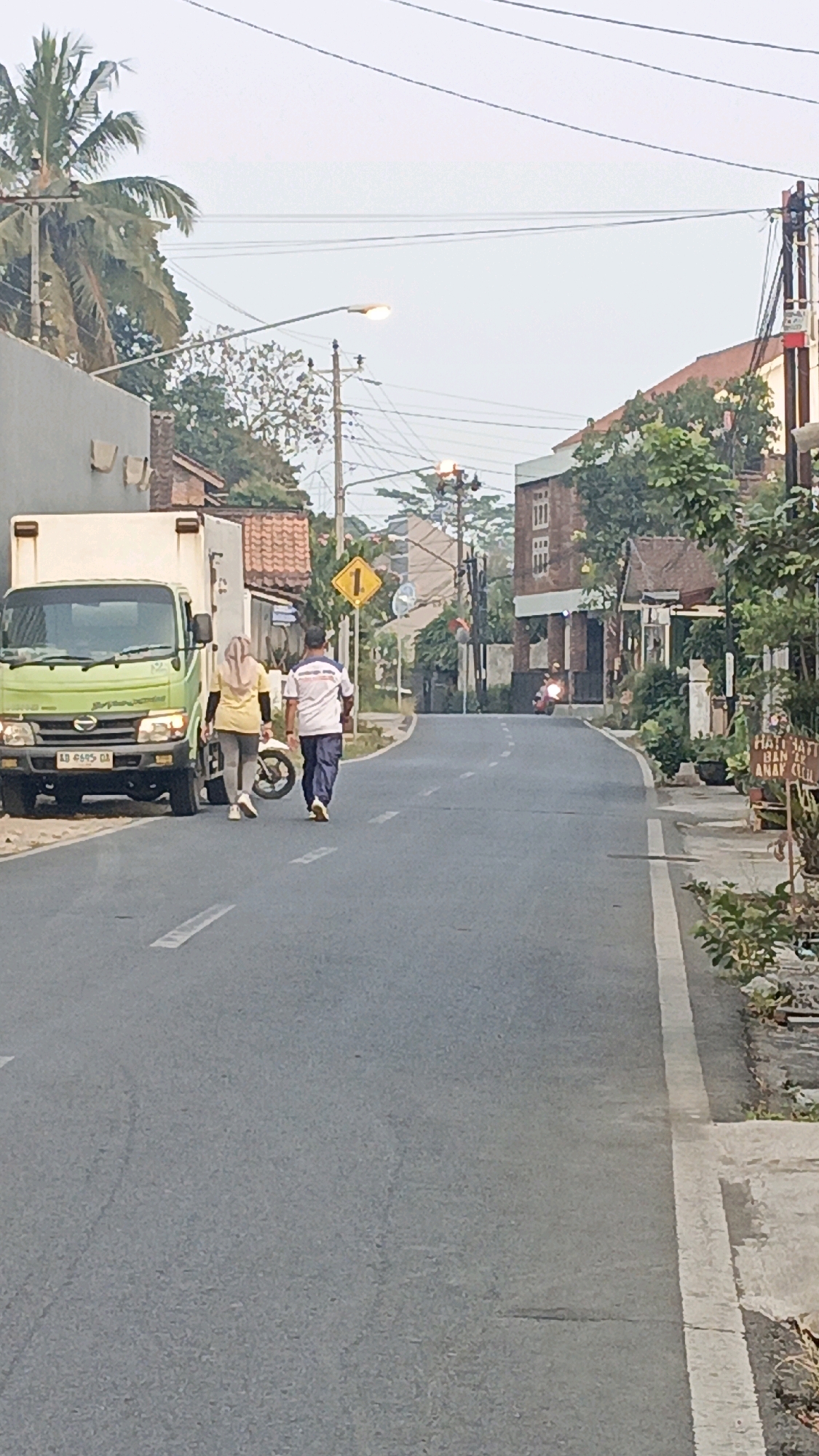 A two-lane asphalt road curves slightly uphill and to the right. A light green truck is parked on the left side of the road. Two individuals, a woman wearing a yellow hijab and a gray top, and a man in a white shirt with blue accents and dark pants, are walking away from the viewer on the left side of the road. A white motorcycle is parked between them and the truck.
Further up the road on the left, a yellow diamond-shaped traffic sign with a black number "1" is visible. On the right side of the road, several buildings are visible, including a two-story structure with brick and white facades. Green trees and plants line the right side of the road. A sign on the right reads "HATI HATI BAN SEREP ANAK KECIL".
In the distance, another person on a motorcycle is traveling uphill. The sky is overcast, suggesting it is early morning or late afternoon. The overall environment appears to be a residential or semi-commercial area within a town or city. The image quality is slightly blurry and pixelated, indicating it was taken with a mobile phone camera. Based on the license plate of the truck, which reads "AD 8495 GA", and the style of the buildings and vegetation, this scene is likely located in Salatiga, Indonesia.