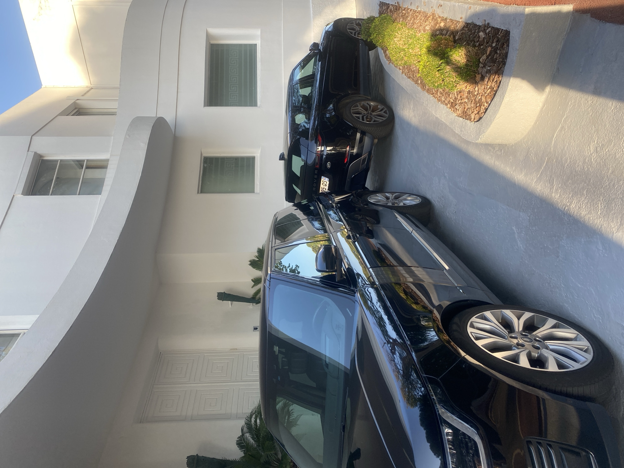 The image depicts the exterior of a modern white building, possibly a residence, located in Antibes, France. Two black Range Rover SUVs are parked in front of the building. The vehicle on the left is a larger model, partially visible from the front quarter, while the vehicle on the right is a smaller model, seen from the rear quarter. The building features curved architectural elements and several windows with decorative grilles. The ground is paved, and a small landscaped area with green bushes and mulch is visible beside the right-hand vehicle. Sunlight casts shadows across the scene, indicating it is daytime. The overall impression is one of luxury and contemporary design.