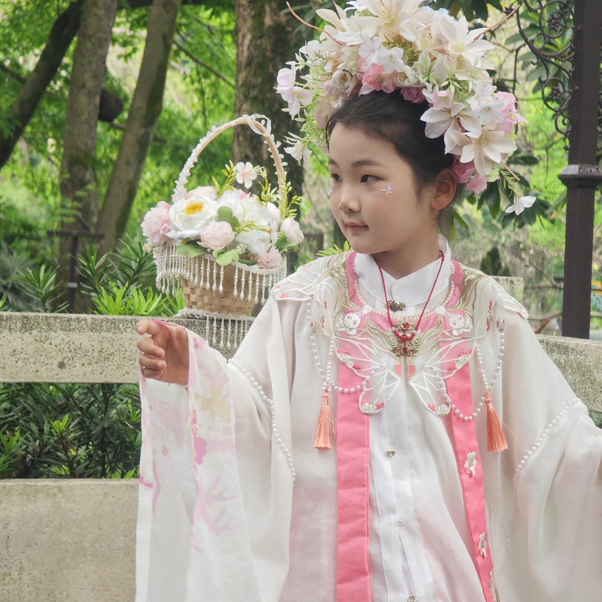 A young girl in a traditional white and pink Chinese dress and a large flower crown is standing near a low stone wall with a flower basket next to her. She is looking to her right. The background features lush green trees and vegetation, and a decorative wrought iron post. The location is likely in a park or garden. The city is Yuzhong District, China.