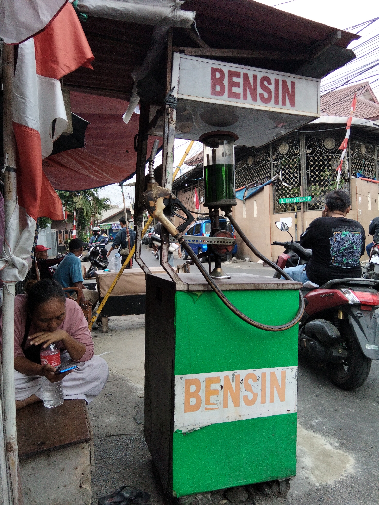 The image depicts a roadside gas station in East Jakarta, Indonesia. The primary focus is a green gas pump with a glass cylinder filled with green liquid, and a nozzle attached by a hose. Above the pump, a sign reads "BENSIN" in red letters. To the left, a woman in a pink shirt and white pants sits on a wooden box, holding a bottle of water and a blue cellphone. Several Indonesian flags, red and white, are visible in the background, along with a building with intricate metal fencing. A motorcycle rider is seen from behind on a red scooter to the right. Other individuals and vehicles are present in the street, indicating a busy urban environment. The ground is paved with asphalt and gravel. The overall scene suggests a typical street view of a local fuel vendor.
