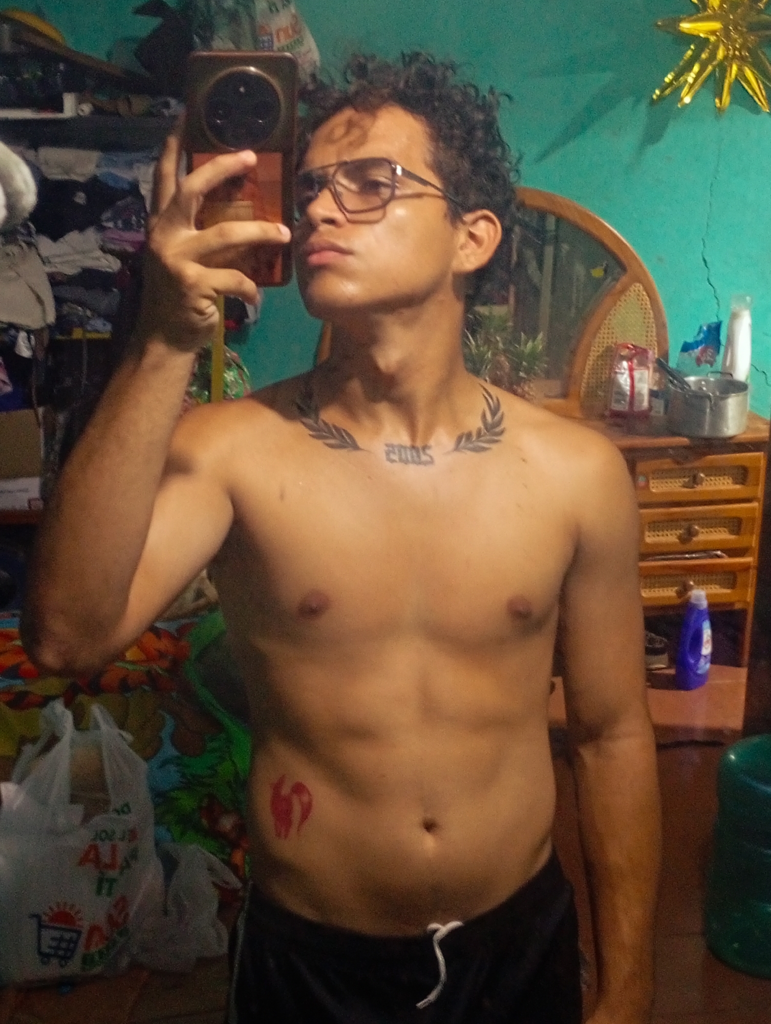 A young, shirtless man with curly dark hair and glasses is taking a selfie in what appears to be a cluttered room in Altagracia, Venezuela. He is wearing dark shorts with a white drawstring. He has a tattoo of a laurel wreath and the year "2005" on his chest, and another red tattoo on his right side, just above his hip. The background shows shelves filled with various items, a mirror, a wooden dresser with drawers, and plastic bags on the floor. A golden star decoration is visible on the wall to the right.