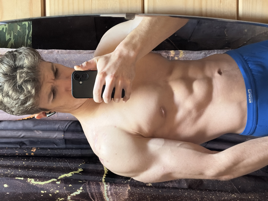 A shirtless man in blue underwear takes a mirror selfie with a black phone. His torso is visible, revealing defined abdominal muscles. He has light skin and curly brown hair. Part of his face is visible, with his eyes closed and brows furrowed. He wears a small earpiece. A mottled black and gold surface is under him. In the background, a camouflage print is visible to the left, and dark and light rectangles can be seen above and to the right.