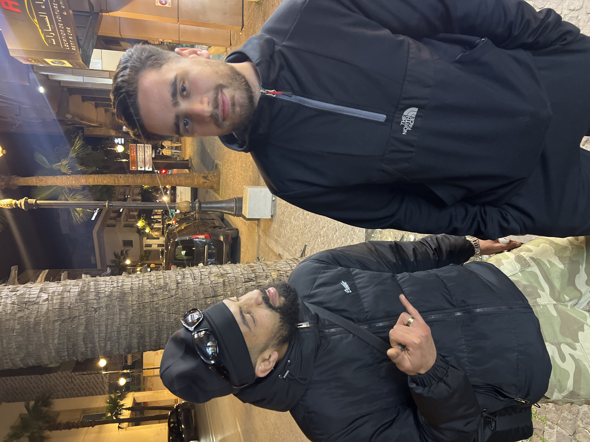 Two men are posing on a street in Tangier, Morocco. The man on the top is wearing a black The North Face jacket. The man below is wearing a black puffer jacket, black beanie, and sunglasses on top of his head. He is gesturing with his right hand, showing his middle finger. Behind them is a street scene with palm trees and buildings. Street signs in Arabic and French can be seen. The man on the top is looking into the camera with a faint smile. The scene is taking place at night, the street is lit up.