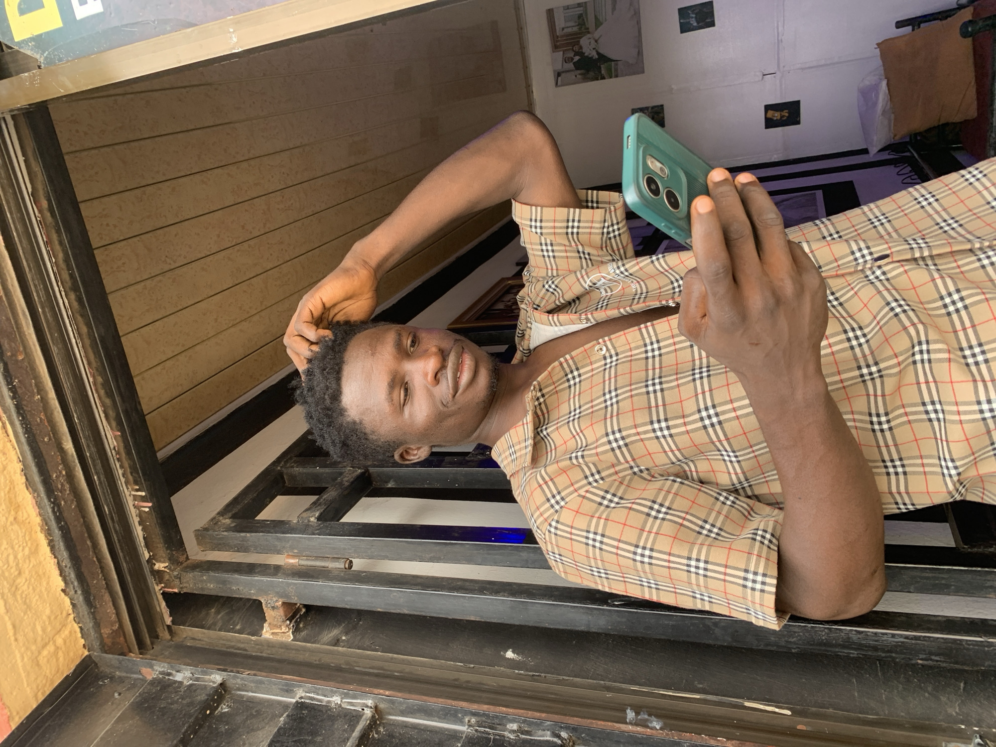 A young African man is seen holding a smartphone in a teal case. He is wearing a plaid button-down shirt and resting his arm on his head. The location appears to be a building interior in Jos, Nigeria, as suggested by the style of architecture and interior design elements. The man is positioned near a window with a black metal frame and a partially visible wooden panel. The background shows a room with personal items such as pictures on the wall and a bed.