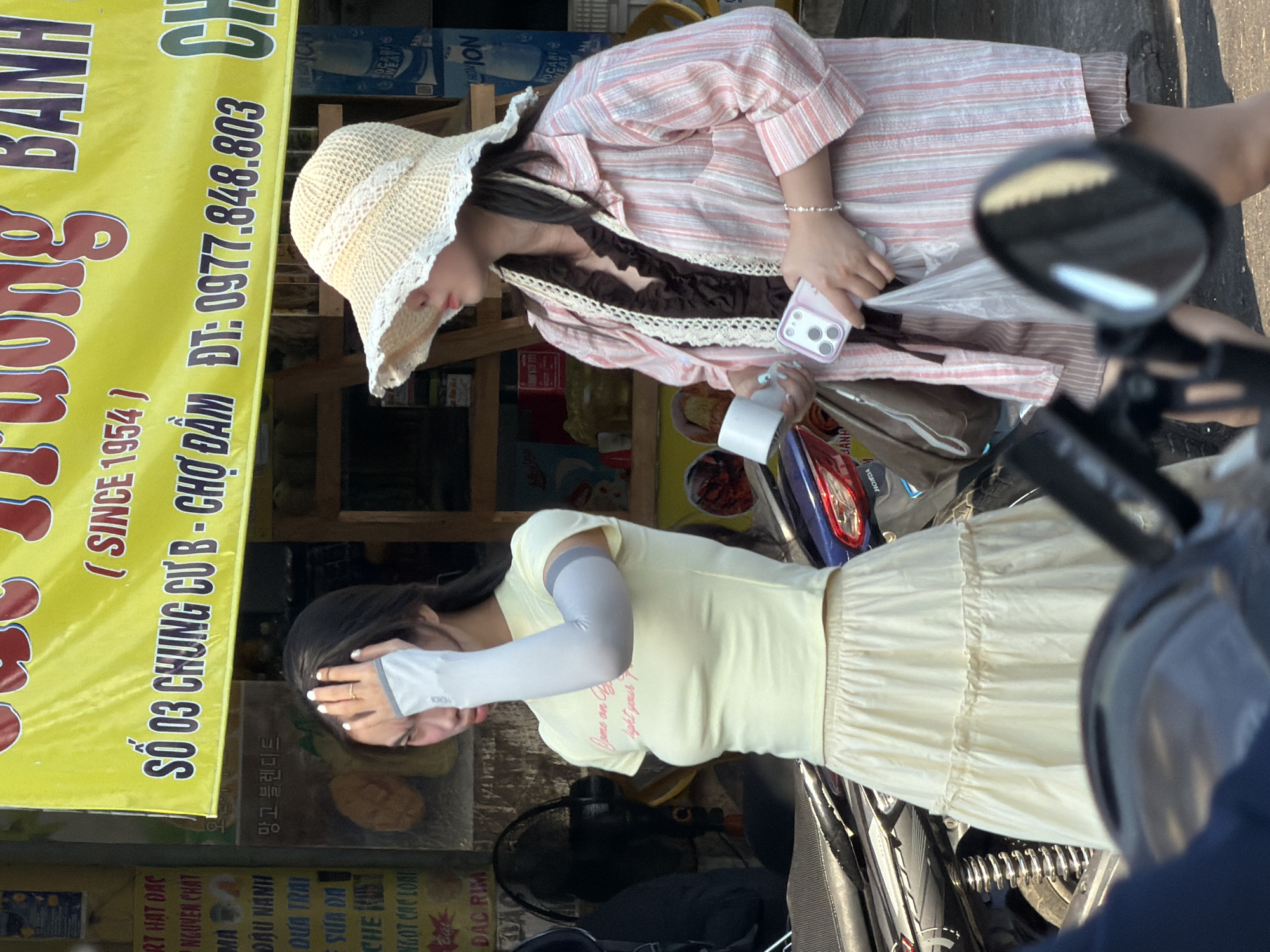 Two young women stand on a city street in Nha Trang, Vietnam. The woman on the left wears a light yellow t-shirt and a cream-colored tiered skirt, with a gray arm sleeve. She has one hand raised to her forehead and appears to be shielding her eyes from the sun. The woman on the right wears a straw hat with a white lace trim, a dark brown ruffled top, and a pink and white striped button-up shirt over a pair of matching striped shorts. She holds a white phone in her right hand and a white cup in her left. Behind them, a bright yellow banner with Vietnamese text is displayed, with the text "SỔ 03 CHUNG CƯ B - CHỢ ĐẦM DT: 0977.848.803" clearly visible. A motorbike is parked between the women, and a black fan is visible to the left. The scene appears to be a street-side market or shop.